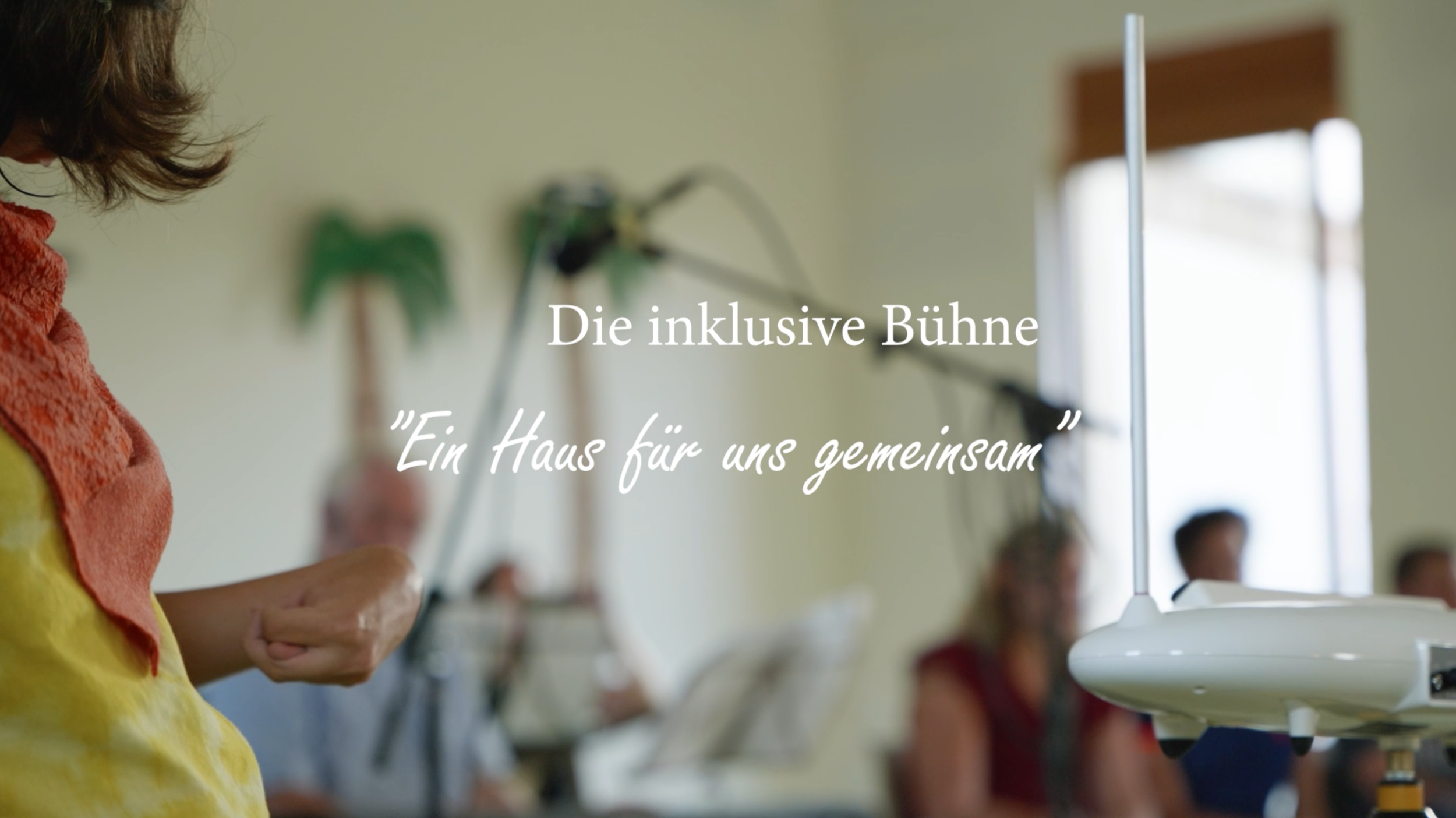 Gallery mit Menschen auf einer inklusiven Bühne, mit Musik-Equipment im Hintergrund, in einem hell erleuchteten Raum. Text auf dem Bild: 'Die inklusive Bühne' und 'Ein Haus für uns gemeinsam'.