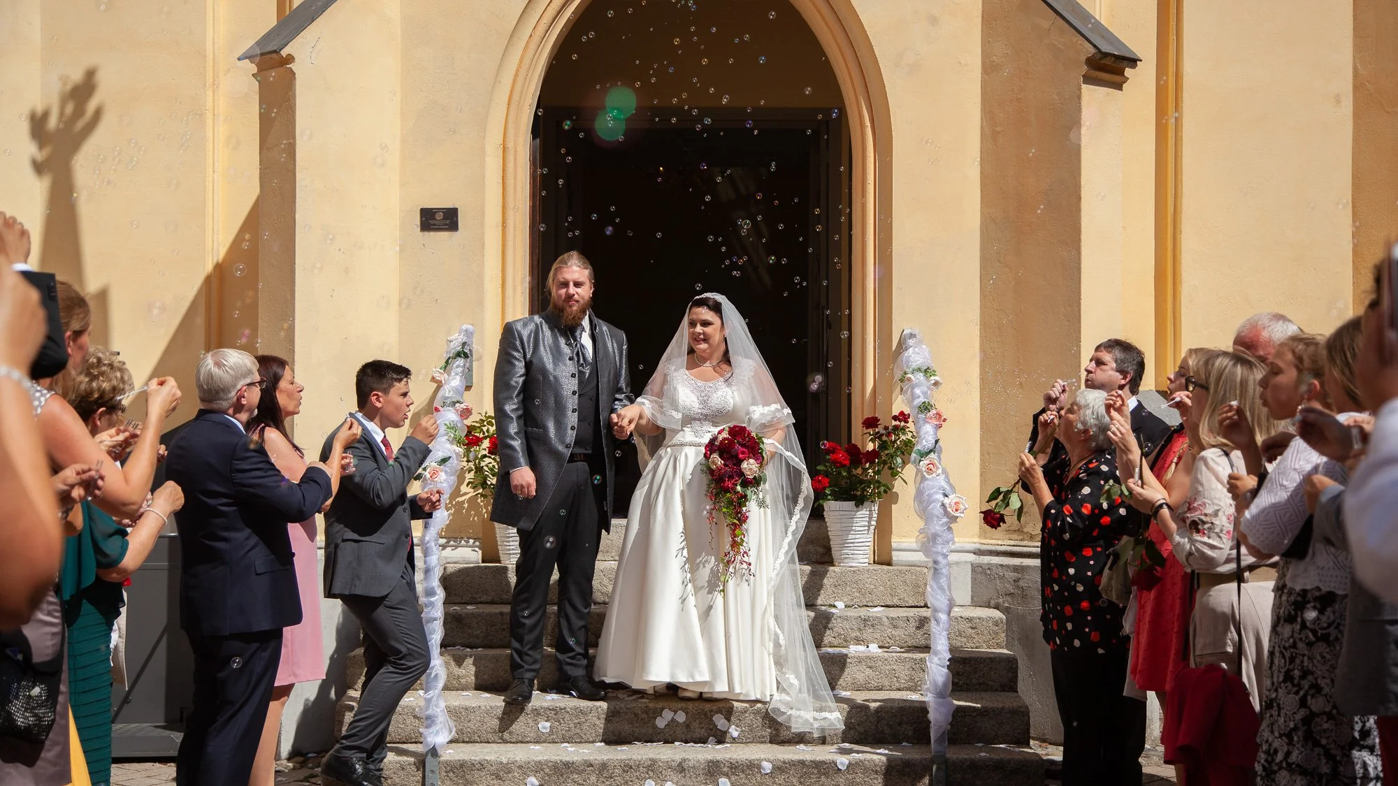 Braut und Bräutigam stehen auf Treppe vor Kirche, umgeben von Hochzeitsgästen, während Seifenblasen fliegen.