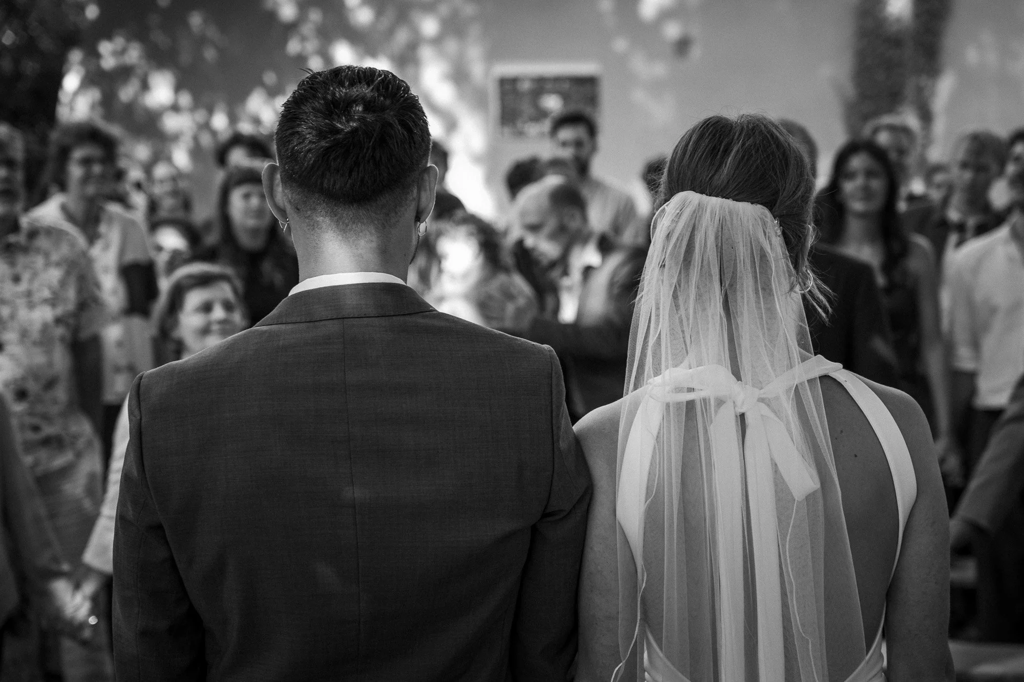 Braut in Hochzeitskleid mit Schleier vor einer versammelten Hochzeitsgäste bei einer Trauung im Freien, Ansicht von hinten.