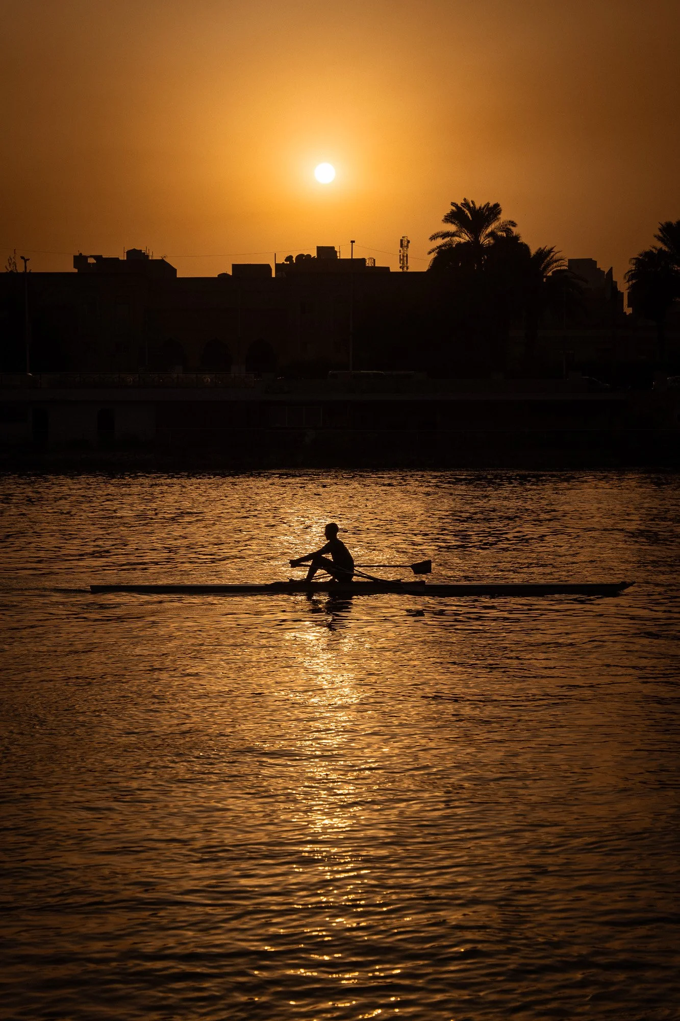 Ein Mensch beim Kajakfahren auf einem Fluss bei Sonnenuntergang, mit Stadt- und Palmen im Hintergrund.