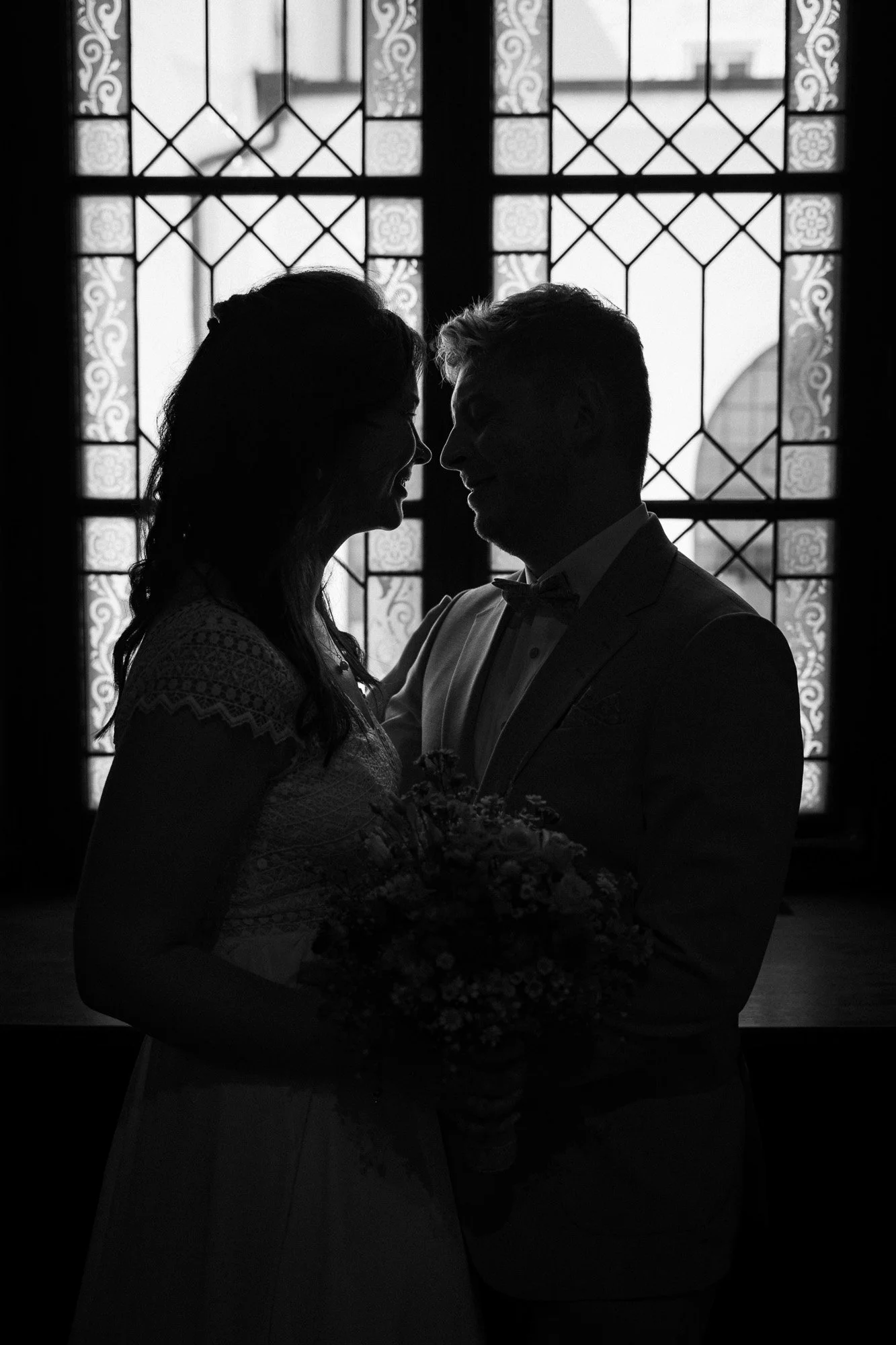 Silhouette eines Paars auf einer Hochzeit vor einem dekorativen Fenster, Frau hält einen Blumenstrauß, Männeranzug mit Fliege, romantische Atmosphäre.