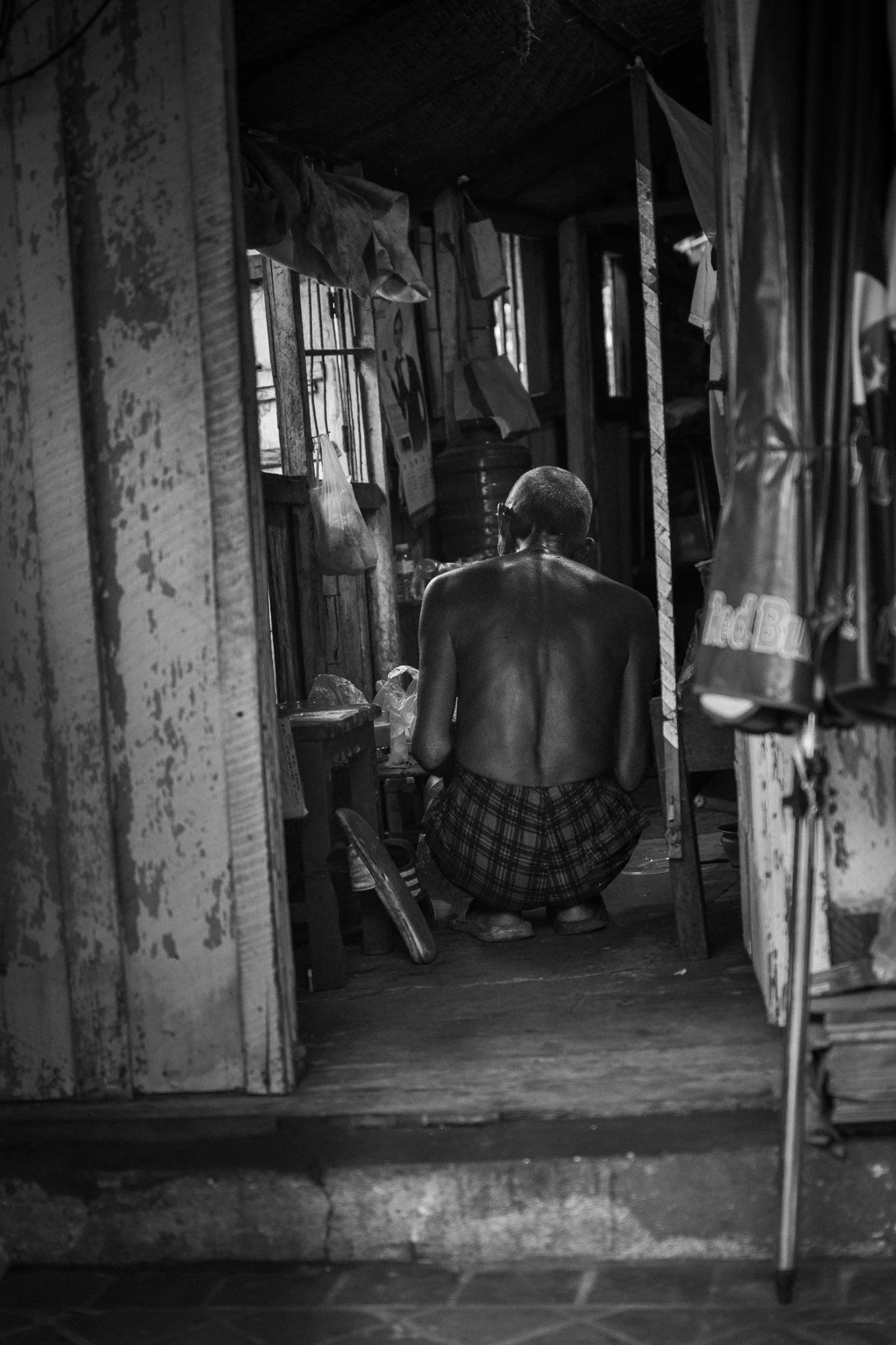 Ein alter, nackter Mann sitzt in einem kleinen, einfachen Raum auf dem Boden, umgeben von alten Möbeln und Gegenständen, in einer ländlichen oder ärmlichen Umgebung, in Schwarz-Weiß-Fotografie aufgenommen.