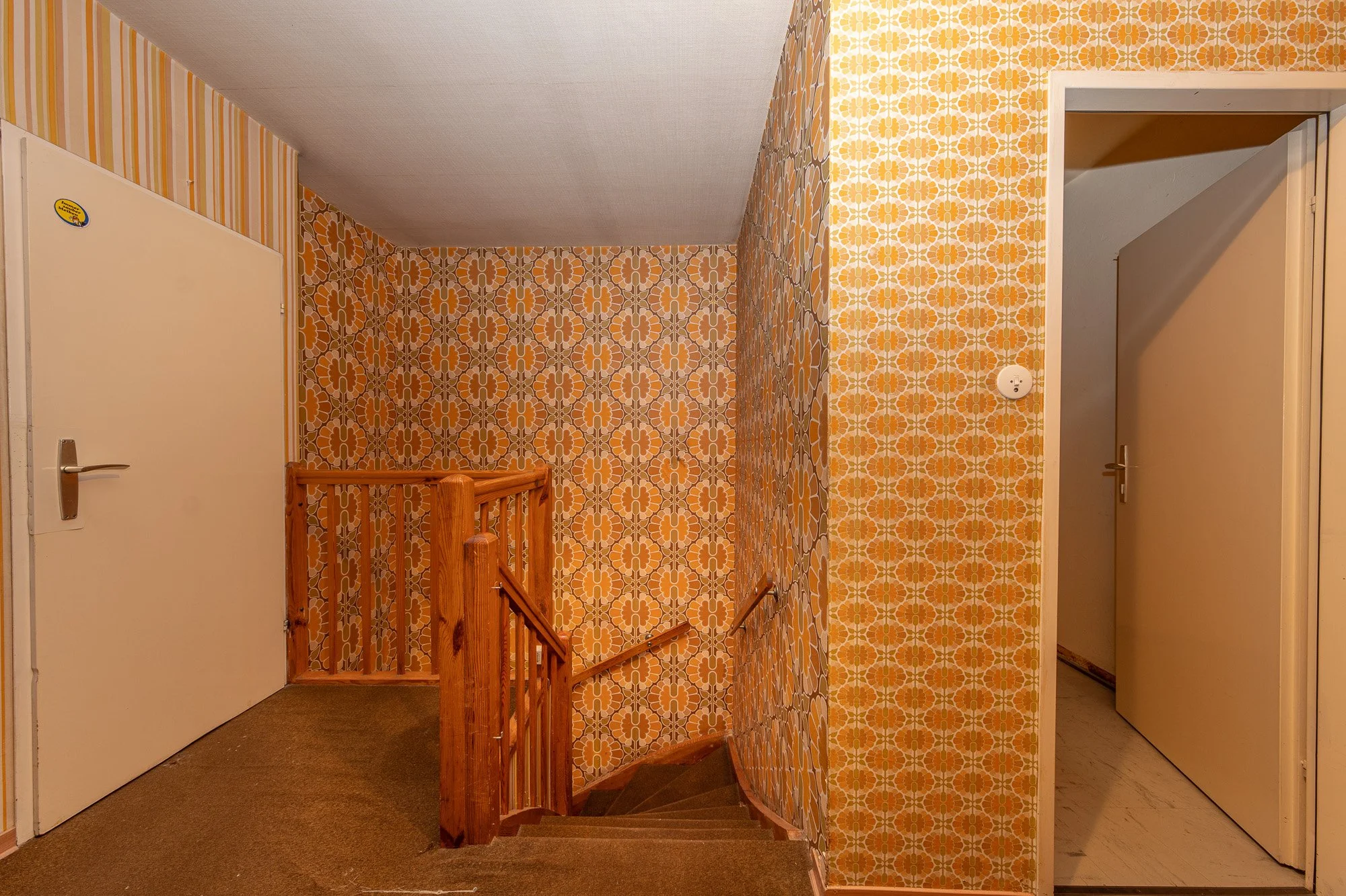 Innenansicht eines Treppenhauses mit orange-brauner Retro- Tapete, Holzgeländer und weißen Türen.