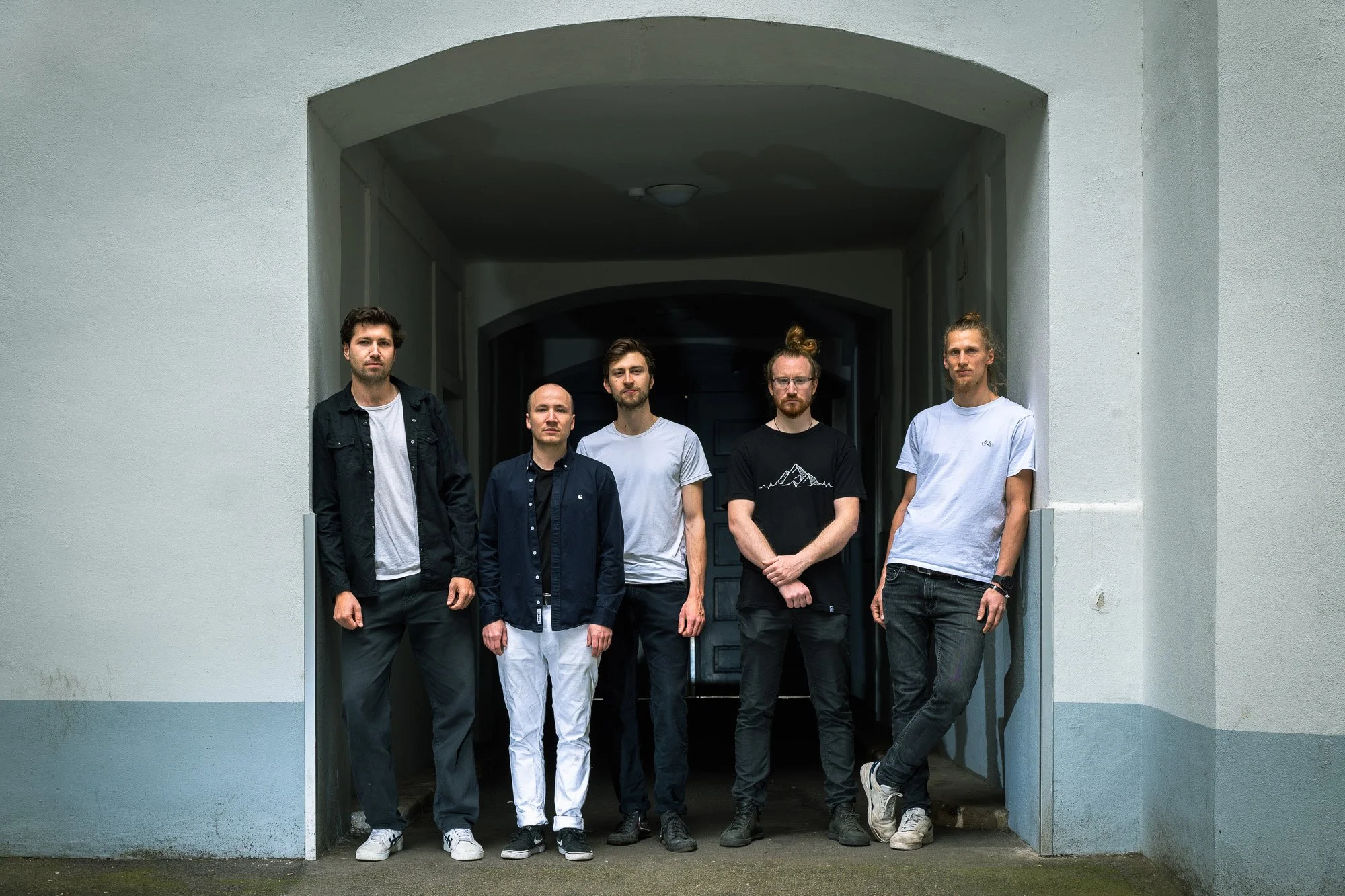 Fünf junge Männer stehen nebeneinander in einem Durchgang vor einer weißen Wand, einige mit Locken oder glatten Haaren, tragen Alltagskleidung und schauen in die Kamera.