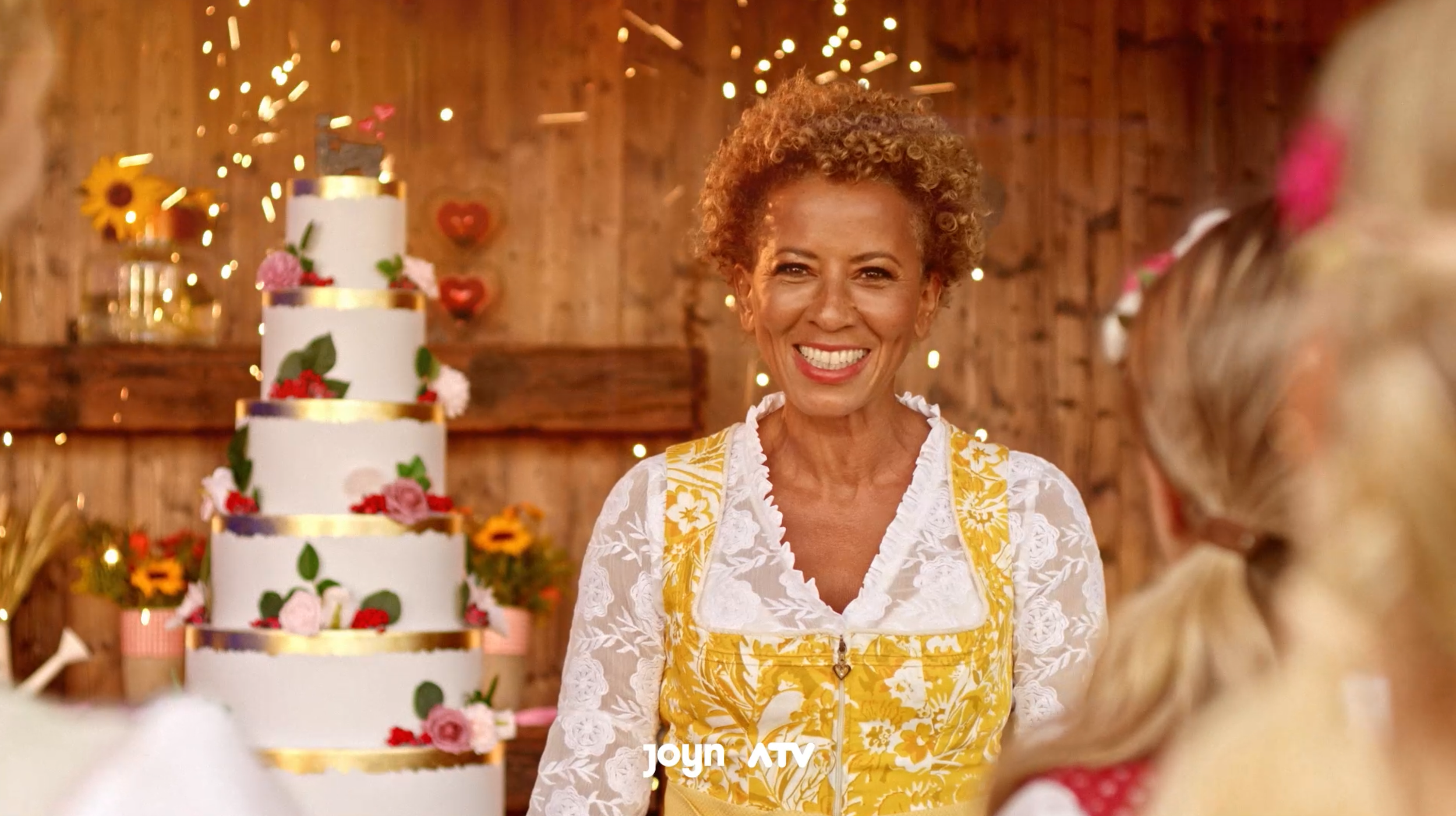 Lächelnde ältere Frau mit kurzen lockigen Haaren steht vor einer mehrstöckigen festlichen Geburtstagstorte, im Hintergrund gedeckte Holzdekoration und leuchtende Lichter. Sie trägt ein weißes Spitzenoberteil mit gelbem Blumenkleid. Im Vordergrund sie