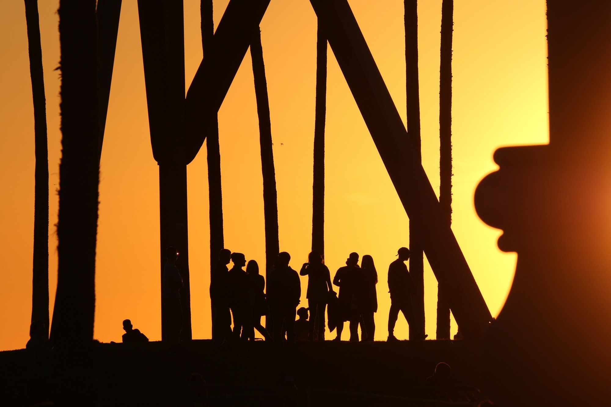 Menschen silhouettiert vor einem orangefarbenen Sonnenuntergang, die auf einer Brücke mit großen Holzpfählen stehen.