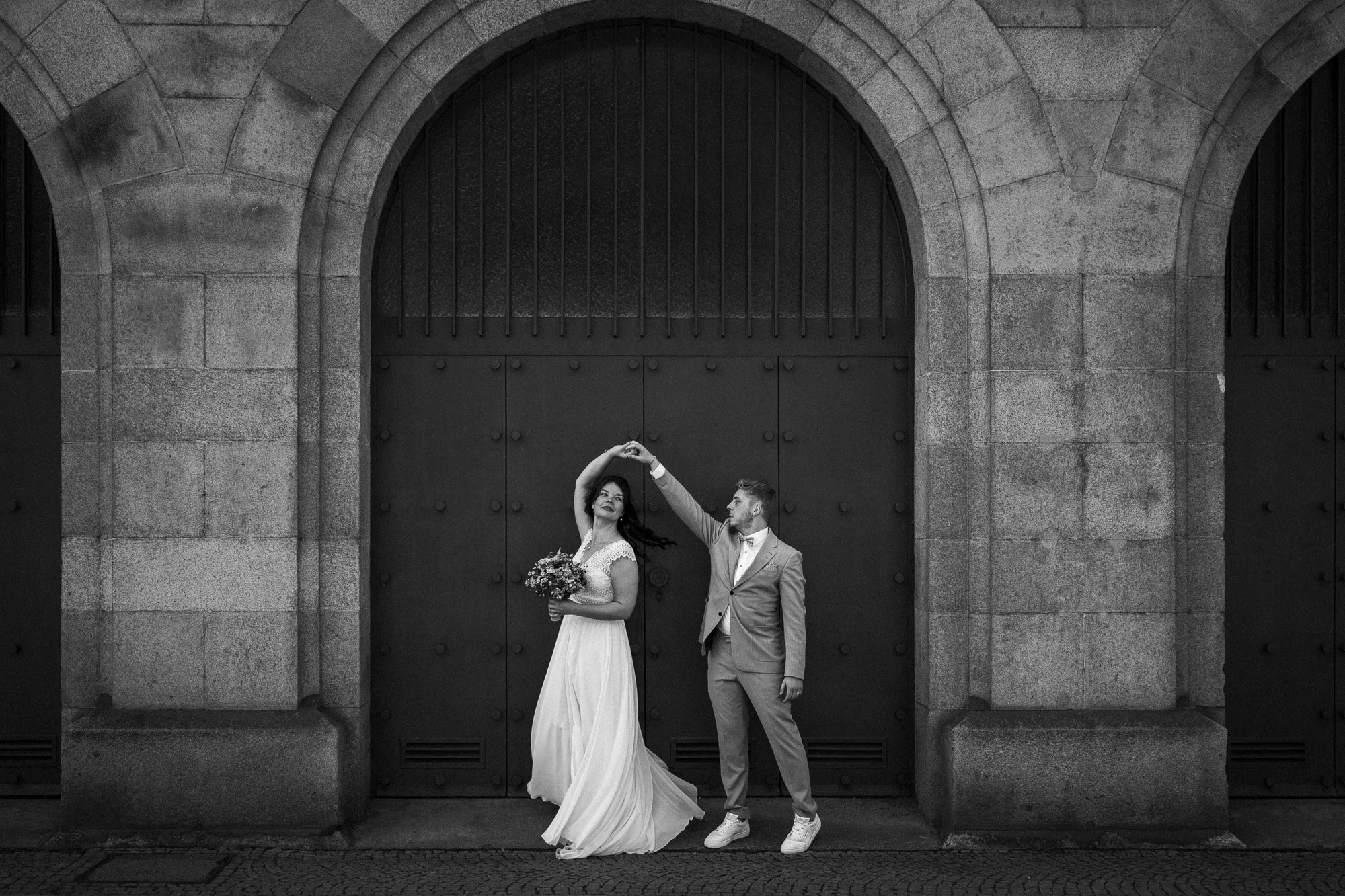 Ein Brautpaar tanzt vor einer großen, alten Steinmauer mit einem großen Tor. Das Bild ist in Schwarz-Weiß.
