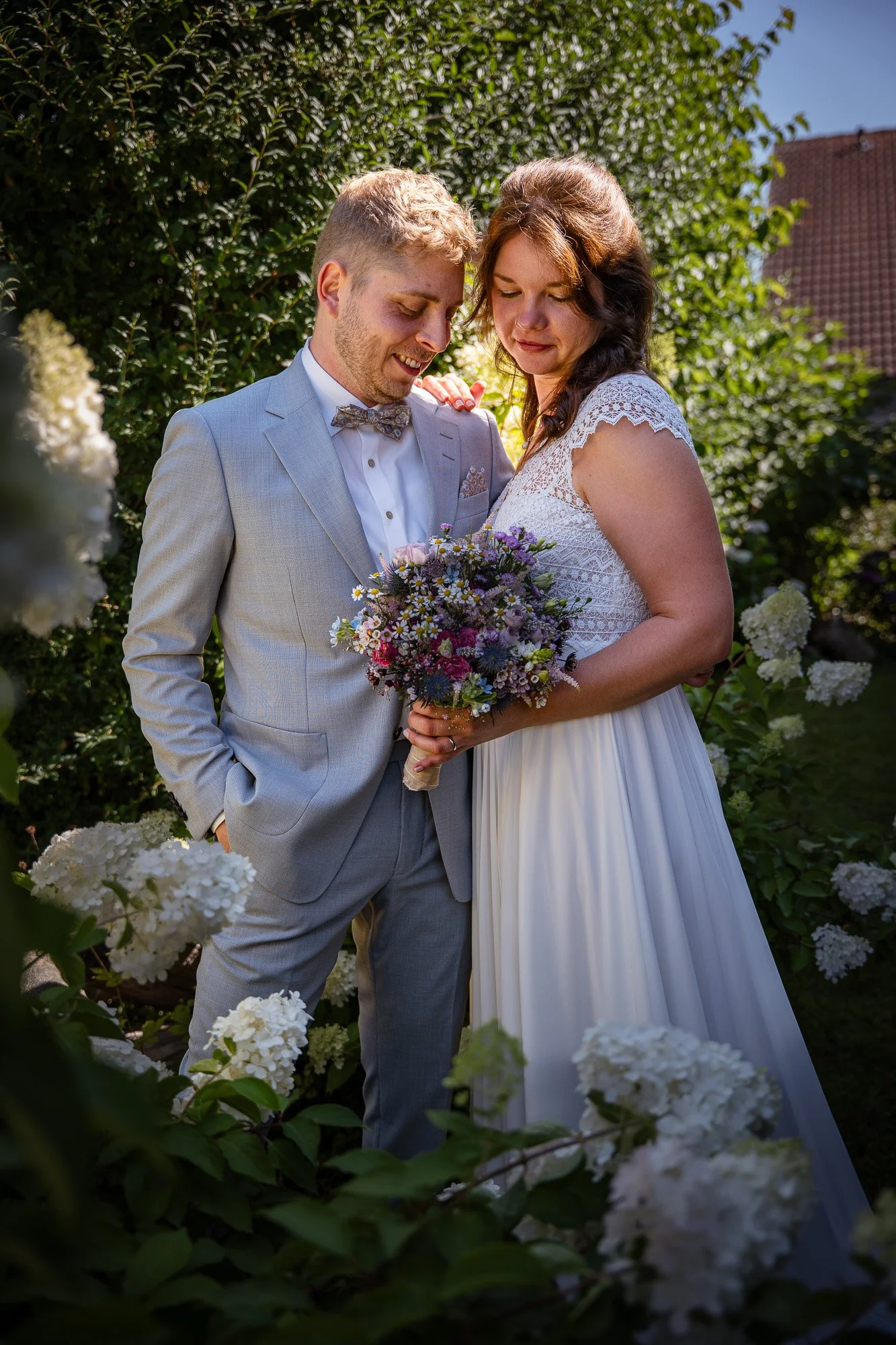 Ein frisch verheiratetes Paar bei einer Hochzeit im Garten, die Frau hält einen bunten Blumenstrauß, beide lächeln und schauen liebevoll auf den Blumenstrauß, umgeben von weiß blühenden Hortensien, sonniger Tag.