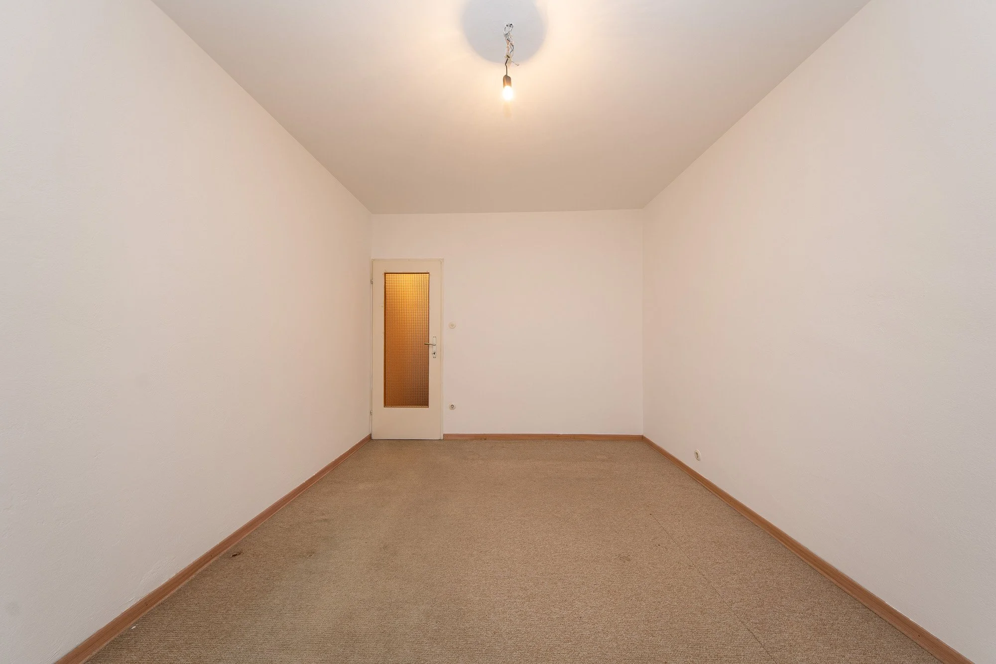 Leeres Zimmer mit weißen Wänden, braunem Teppichboden, einer Tür mit orangem Fenster, und einer einfachen Glühbirne an der Decke.