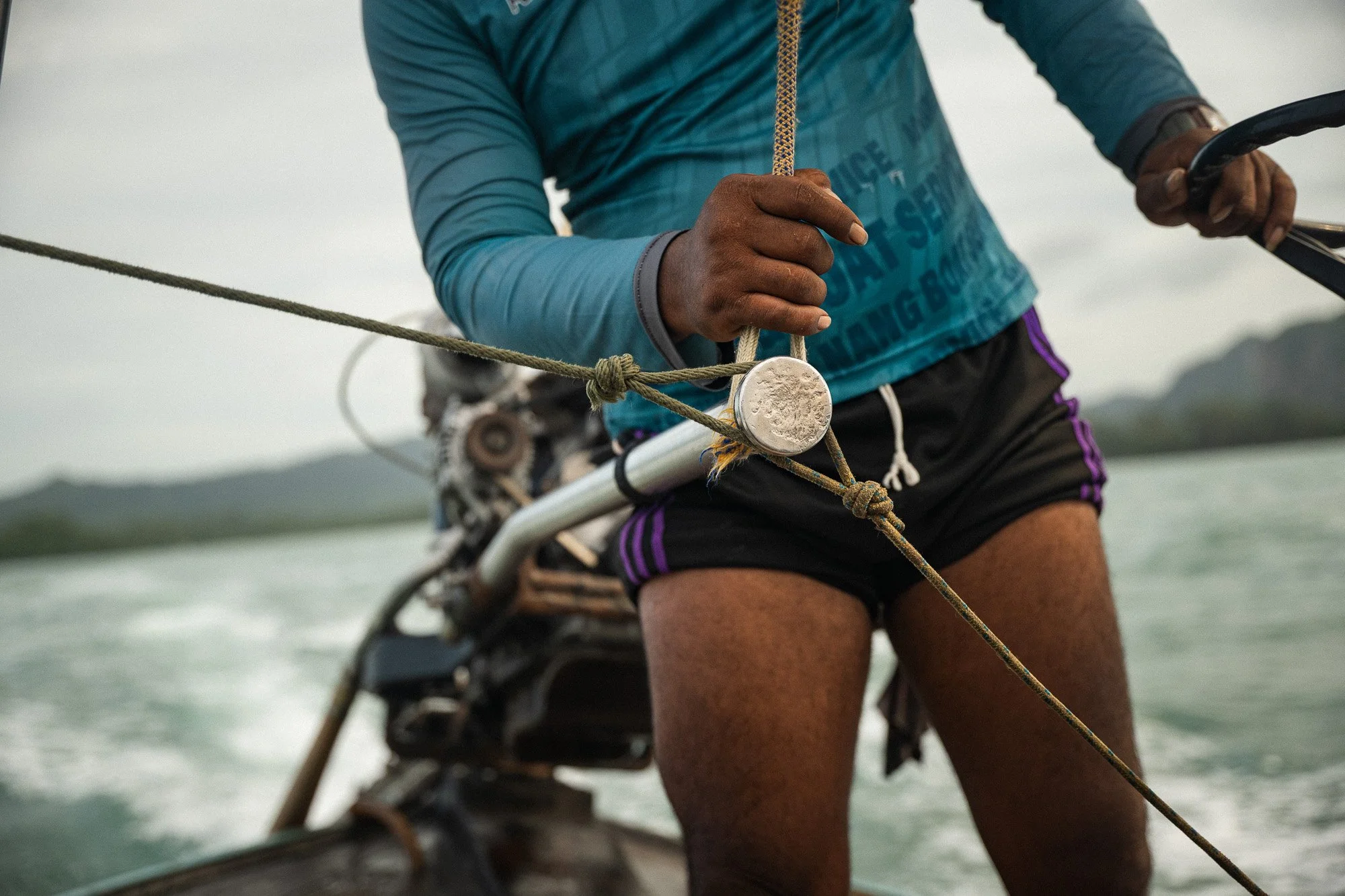 Nahaufnahme eines Menschen, der auf einem Boot segelt, die Hand hält eine Seil und eine Segelklemme, im Hintergrund Wasser und Berge