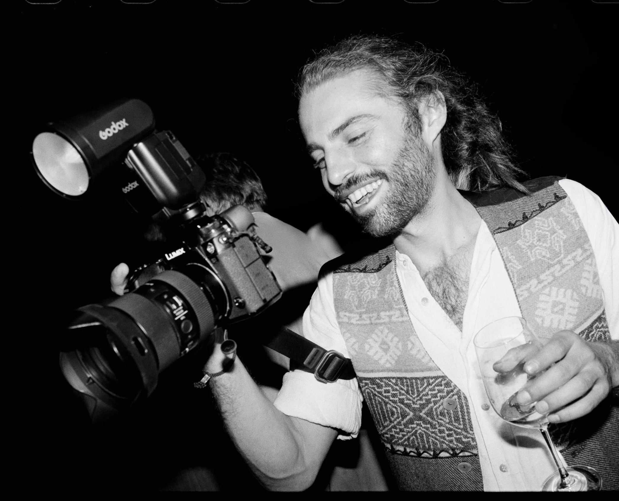 Ein Mann mit langem Haar und Bart, der in einem schwarzen und weißen Bild lächelnd eine Kamera hält und ein Weinglas in der Hand hält.
