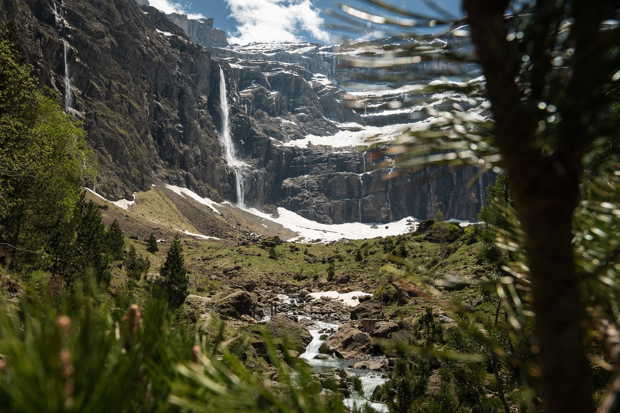 Berglandschaft mit Wasserfällen, Fluss und Tannenbäumen im Vordergrund, Schneeflächen auf den Bergen, bewölkter Himmel