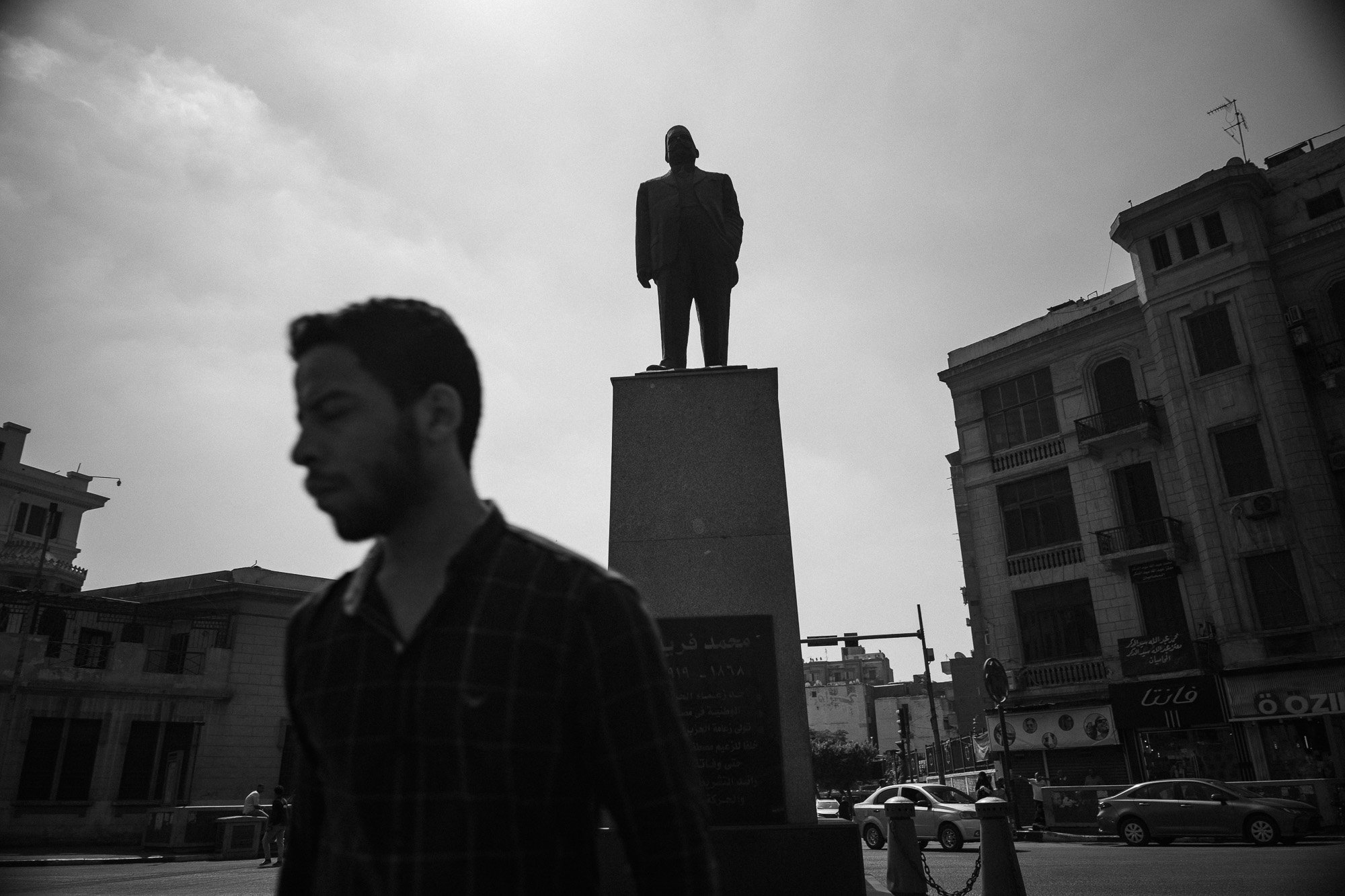 Ein Mann läuft vorbei an einer Statue eines Mannes in Anzügen auf einem Stadtplatz, im Hintergrund sind Gebäude und Autos zu sehen.