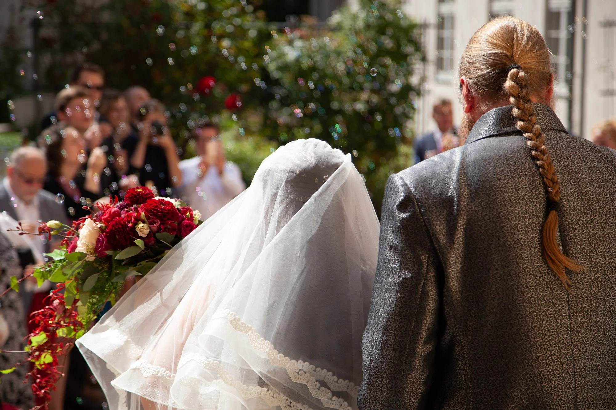 Hochzeitspaar bei der Trauung, Blick auf Braut mit weißem Schleier und Blumenstrauß, Brautvater mit langen geflochtenem Haar, Gäste im Hintergrund, festliche Stimmung, sonniges Wetter