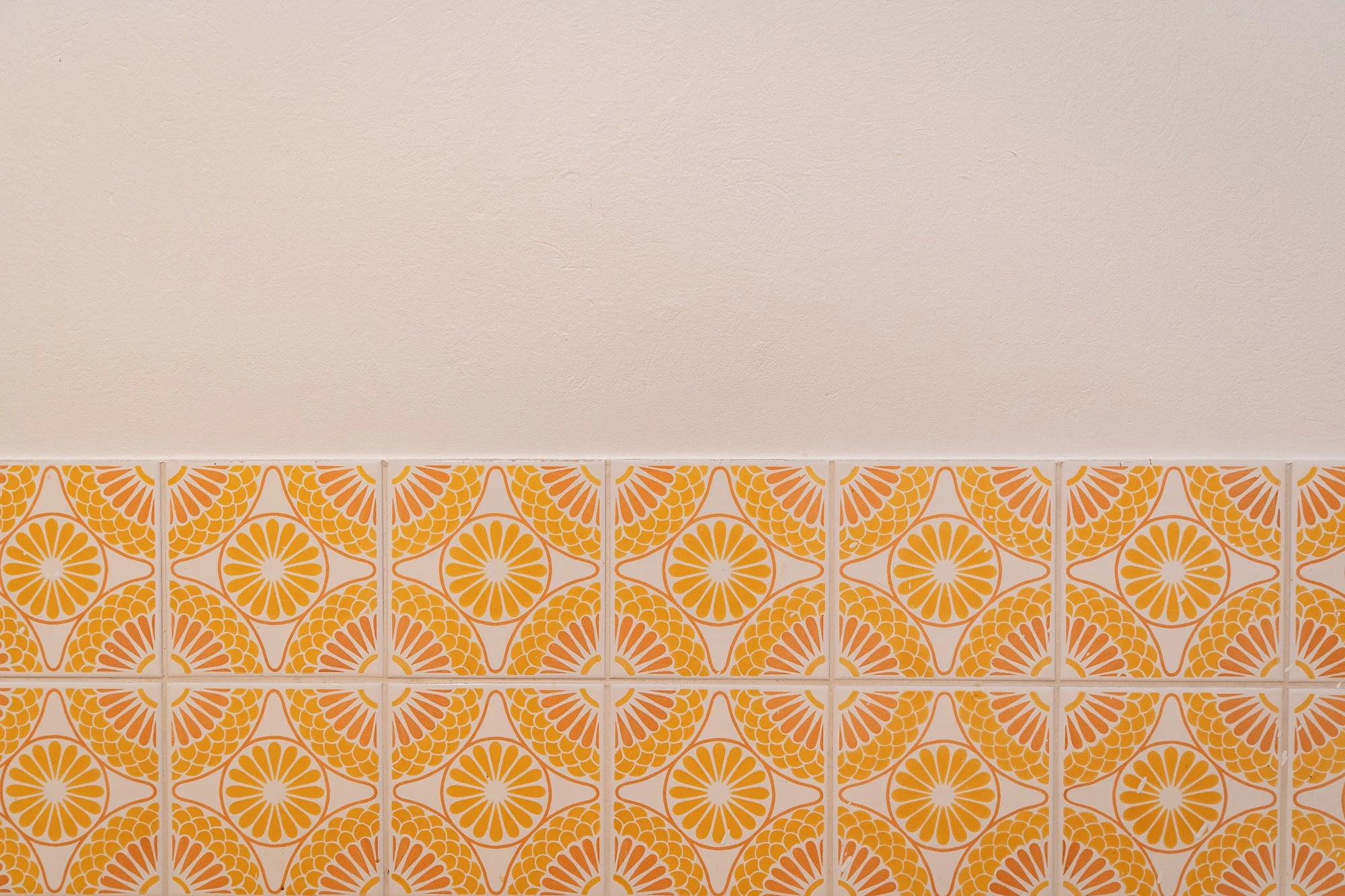 Obere Hälfte der Wand mit glatzer, cremefarbener Oberfläche, untere Hälfte mit orange und weißen geometrischen Fliesen, die ein kreisförmiges Muster bilden.