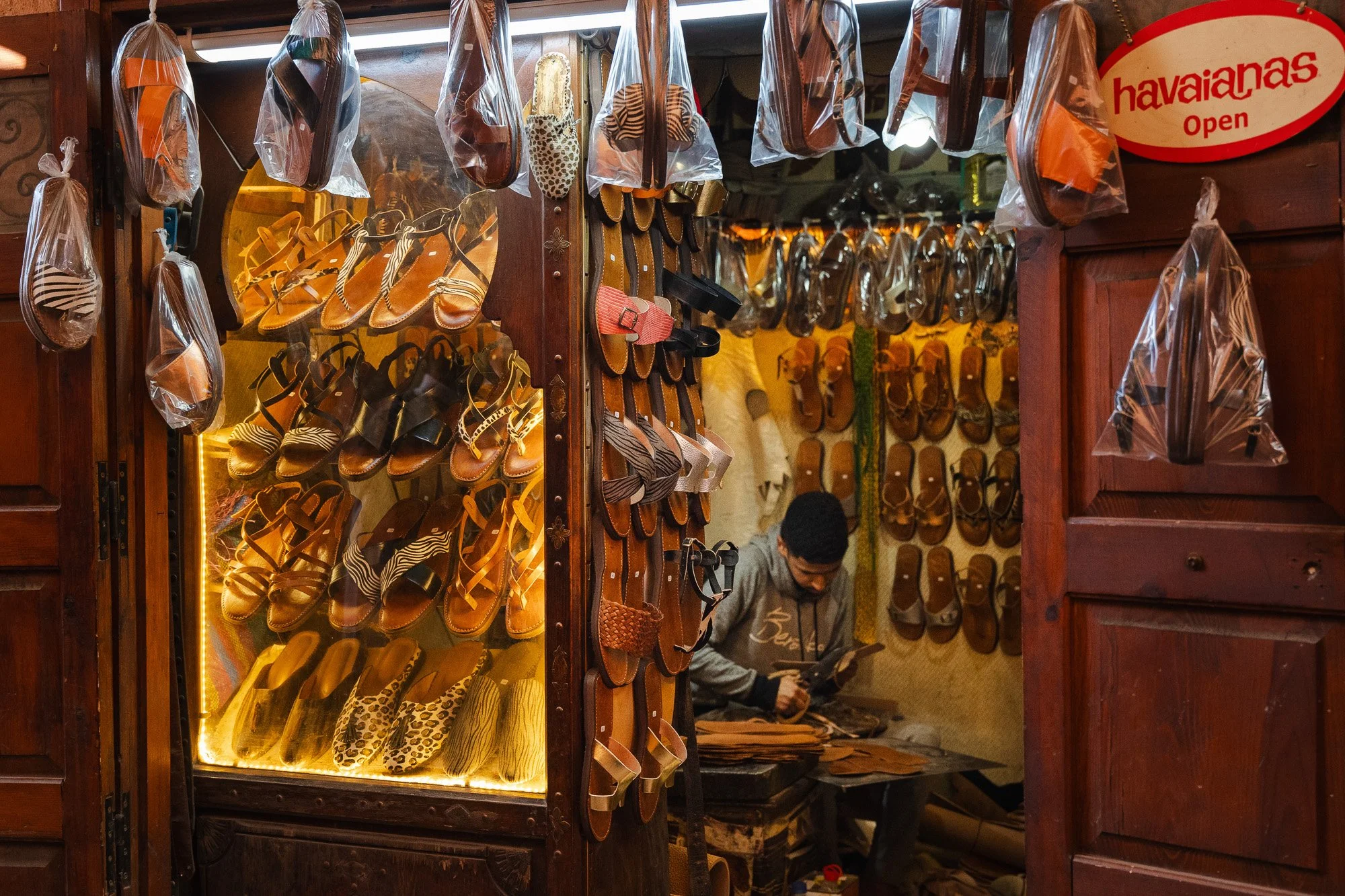 Verkäufer in einem Schuhgeschäft mit Wand voller Sandalen und Flip-Flops, einige in Plastik verpackt, und einer Person, die in der Ecke sitzt und ein Buch liest.