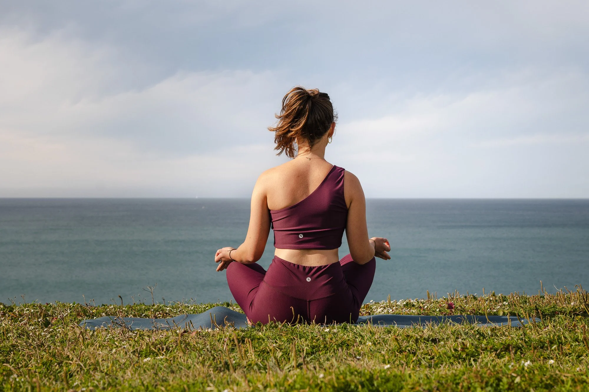 Eine Frau in Sportbekleidung macht Yoga im Sitzen im Freien auf einer Wiese mit Blick auf das Meer, bei bewölktem Himmel.