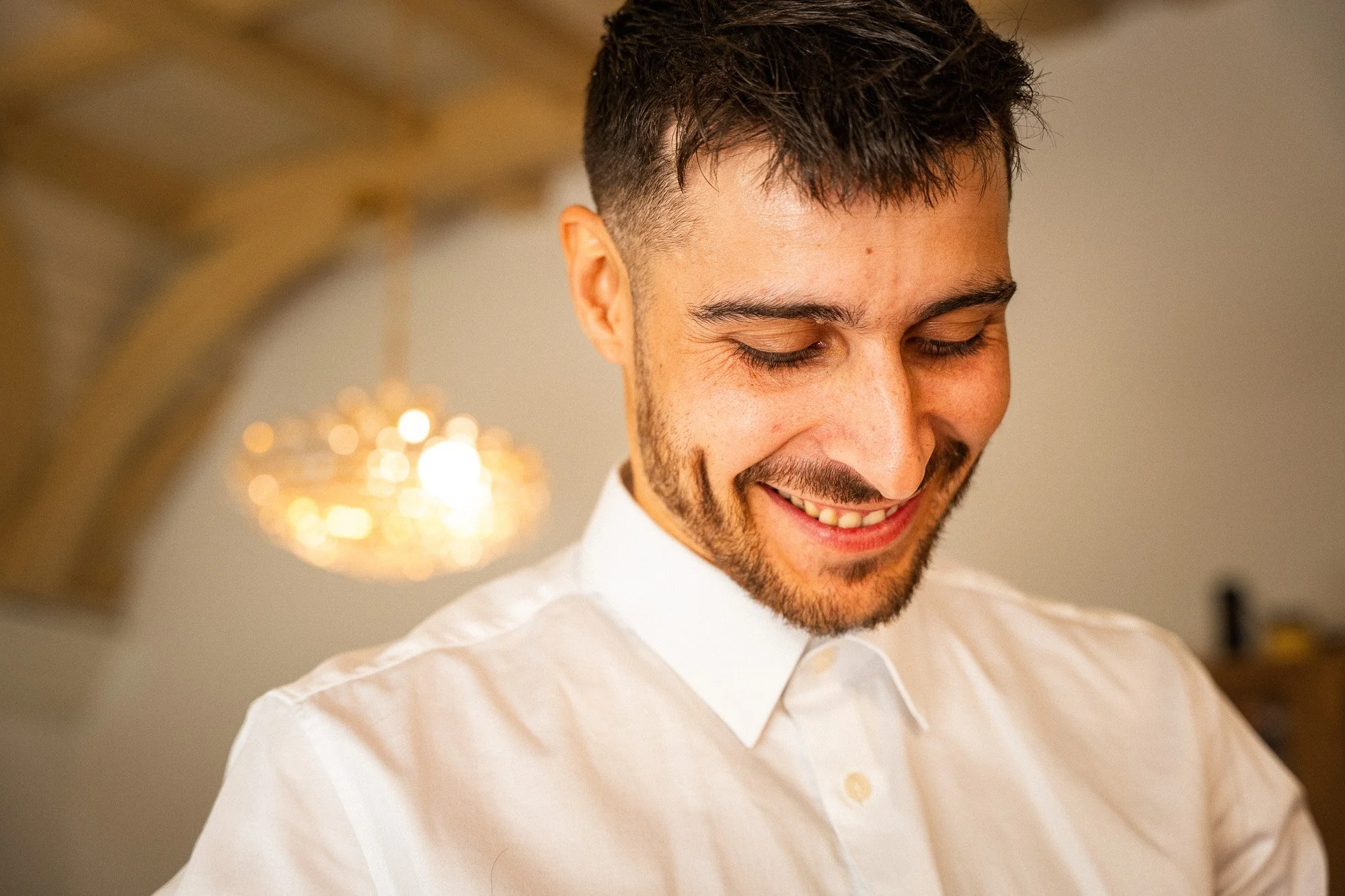Ein lachender Mann in einem weißen Hemd, der nach unten schaut, im Hintergrund eine dekorative Lampe, die warmes Licht ausstrahlt.
