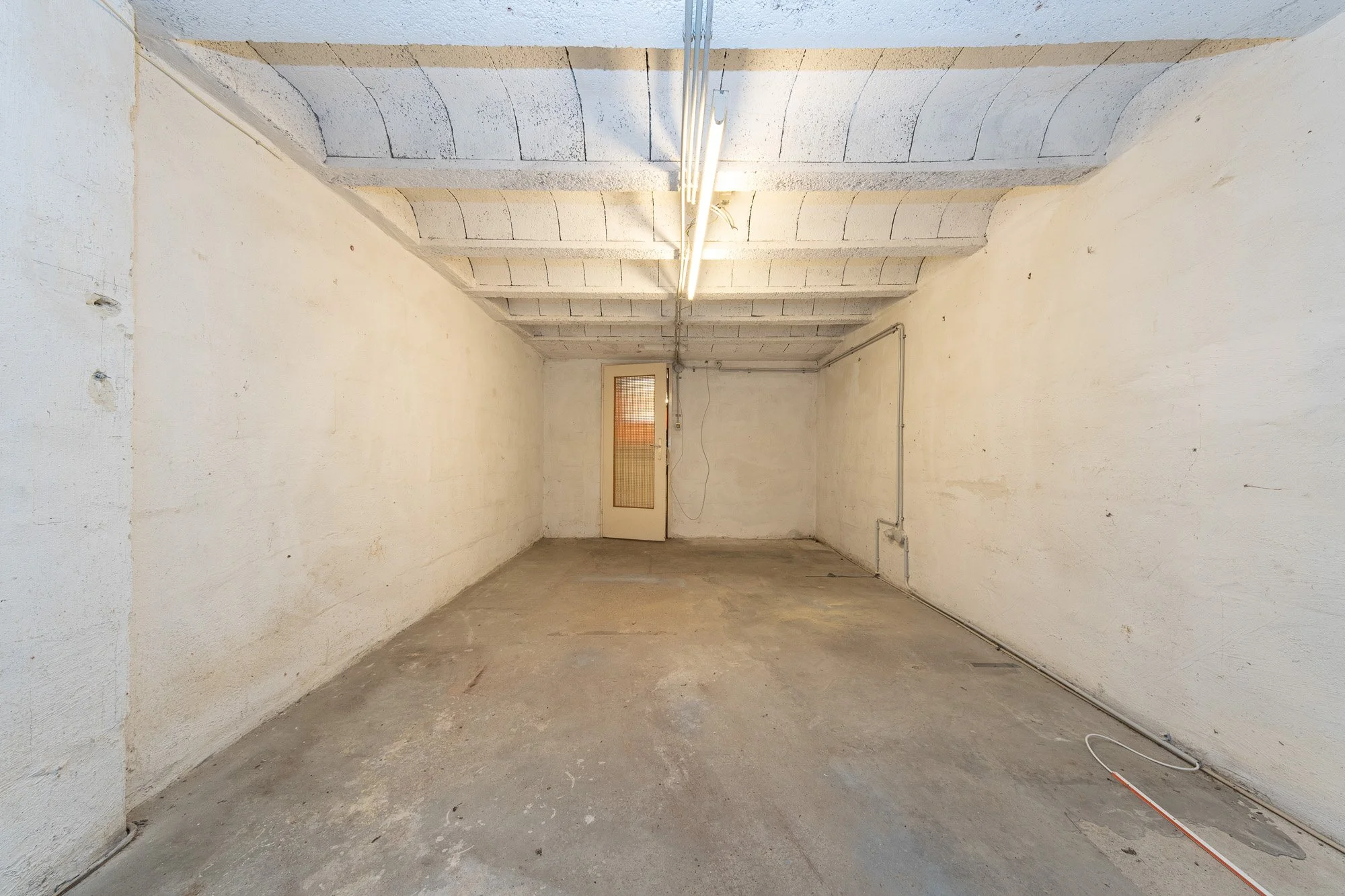 Leeres, unfertiges Garagenraum mit Betonboden, weißen Wänden und Decke, Tür am Ende, elektrische Leitungen an den Wänden und an der Decke.