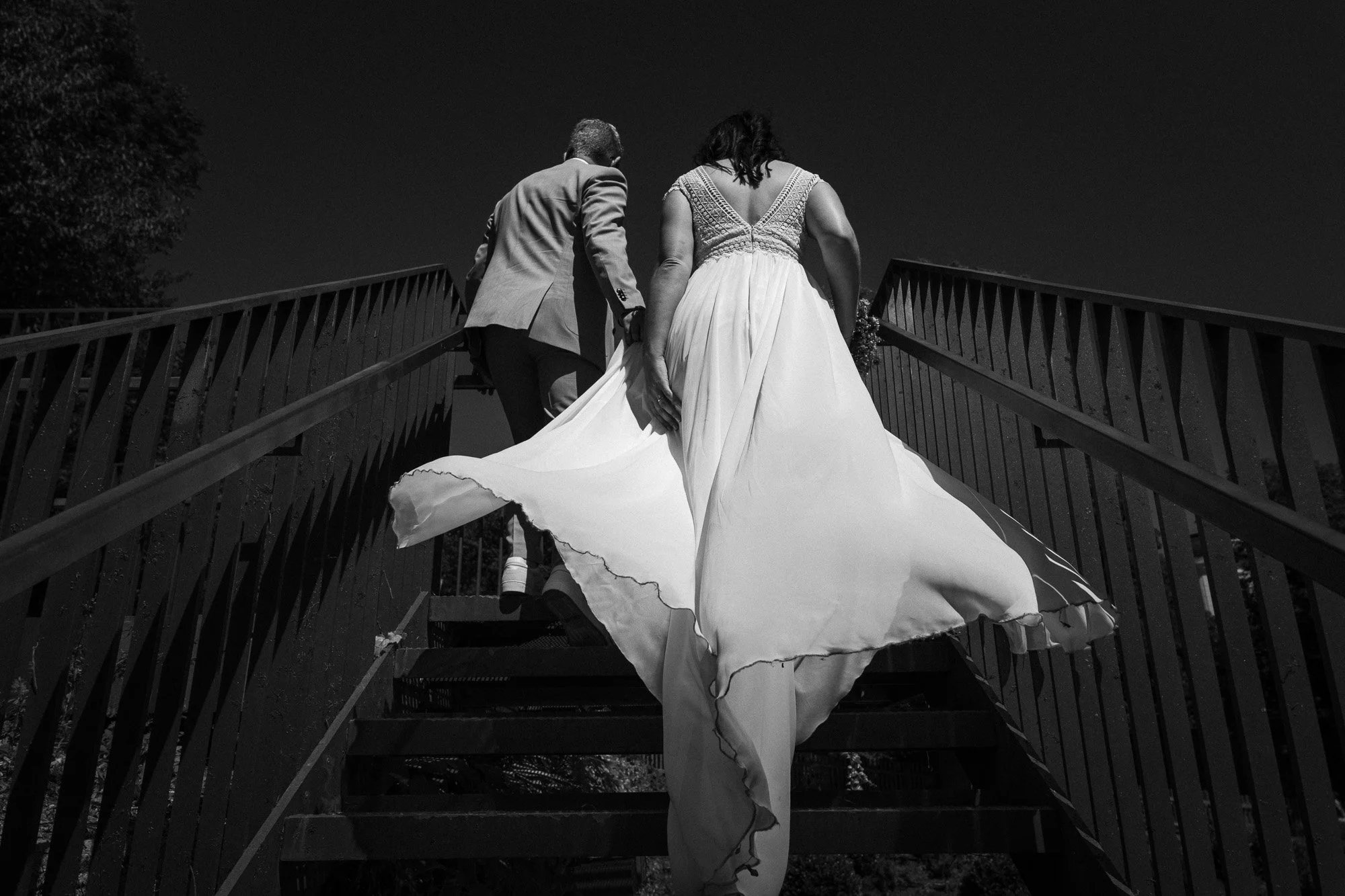 Schwarz-weiß-Foto eines Paares in Braut- und Bräutigamkleidung, das einen Aufstieg auf eine Treppe aus Metall mit Geländern macht, bei Nacht
