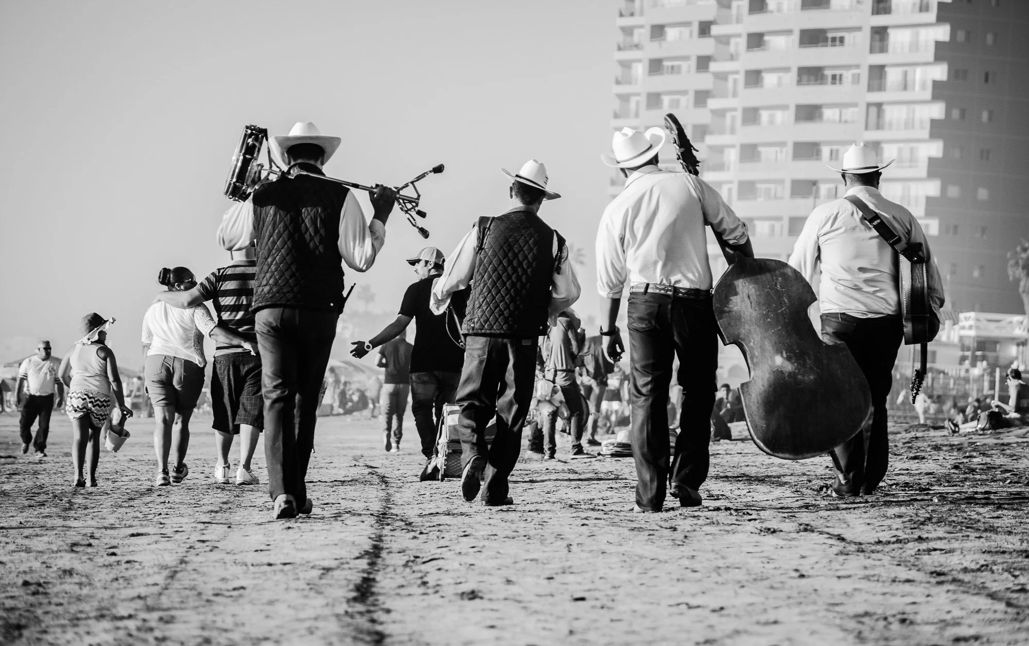 Musiker in Cowboy-Kleidung, die am Strand mit Instrumenten laufen, darunter ein Gitarrist und ein Blasinstrument-Spieler, im Hintergrund Menschen und Hochhäuser.