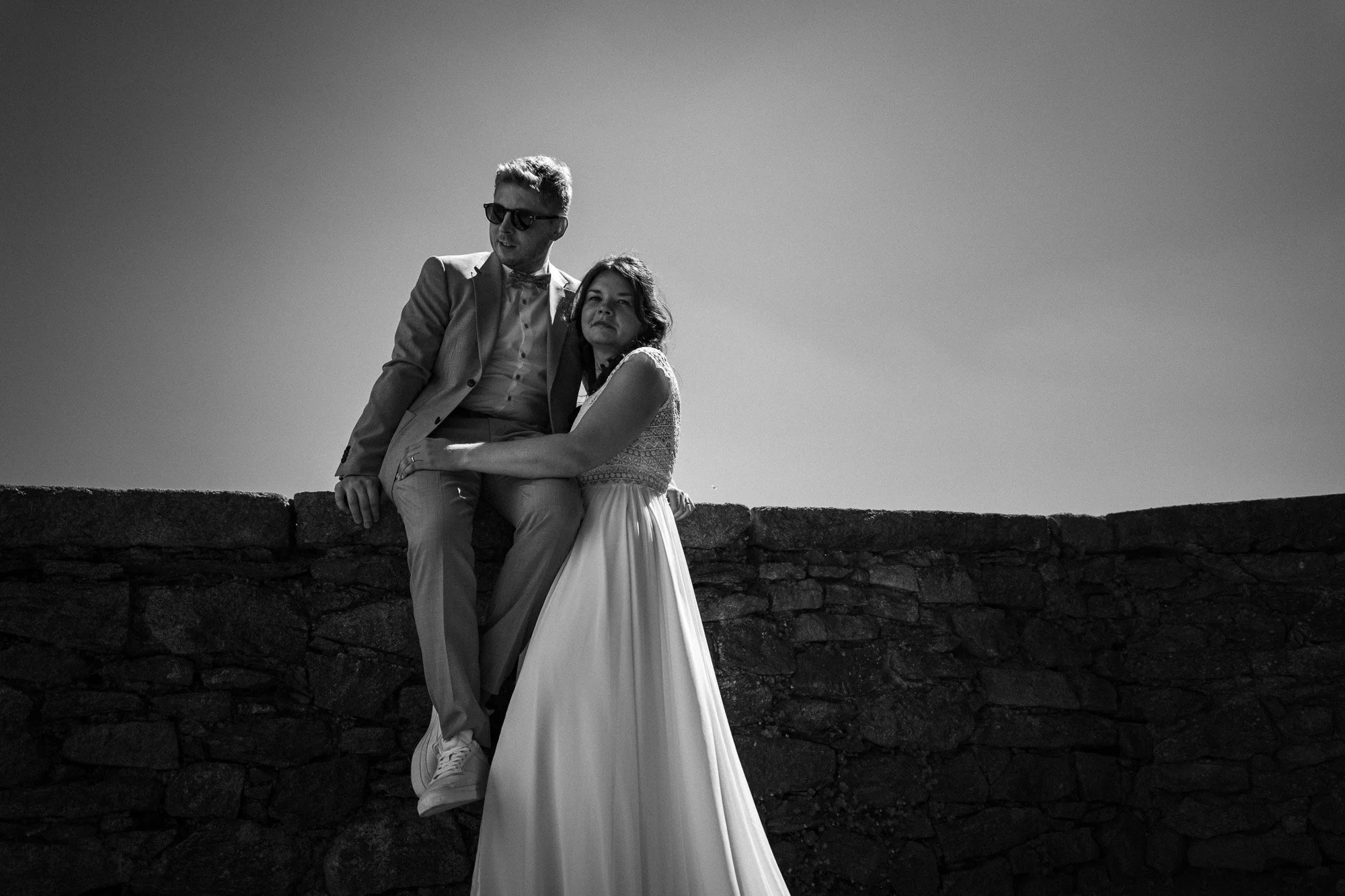 Schwarz-weiß-Foto eines Paares, das auf einer Steinmauer sitzt, bei Sonnenuntergang, romantisch, elegant gekleidet, der Mann trägt Sonnenbrille, Frauen trägt ein langes Kleid.