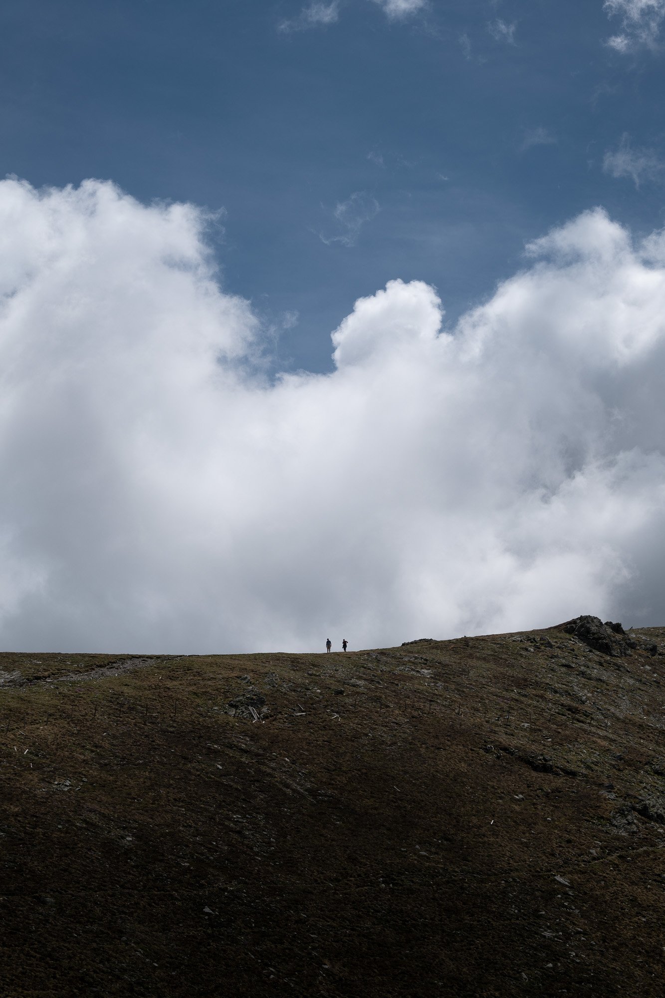 Zwei Personen stehen auf einer hügeligen Landschaft unter einem Himmel mit großen weißen Wolken.