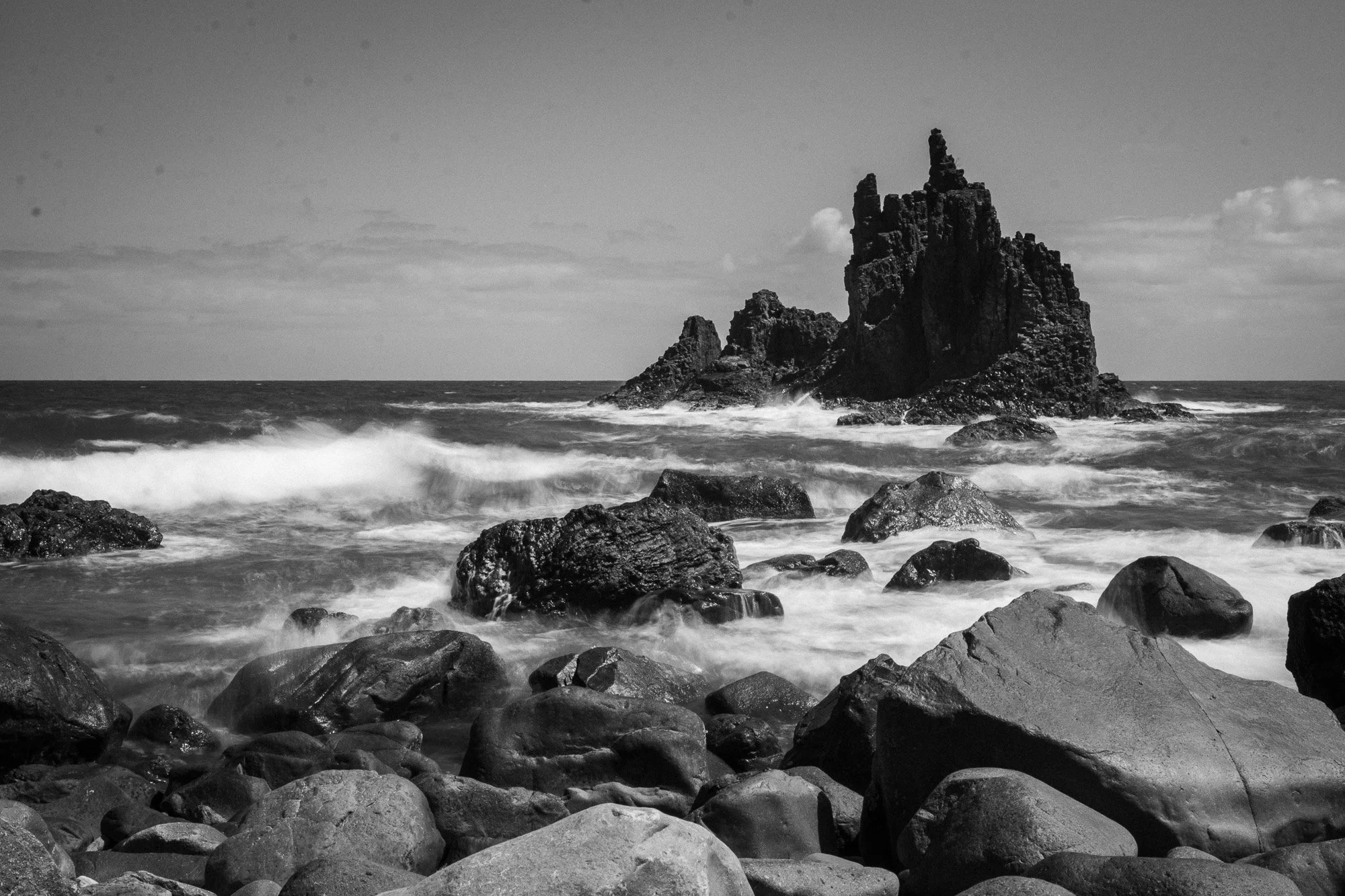 Schwarz-weiß-Bild eines felsigen Meeresufers mit großen Steinen und einem einzelnen hohen Felsen im Wasser.