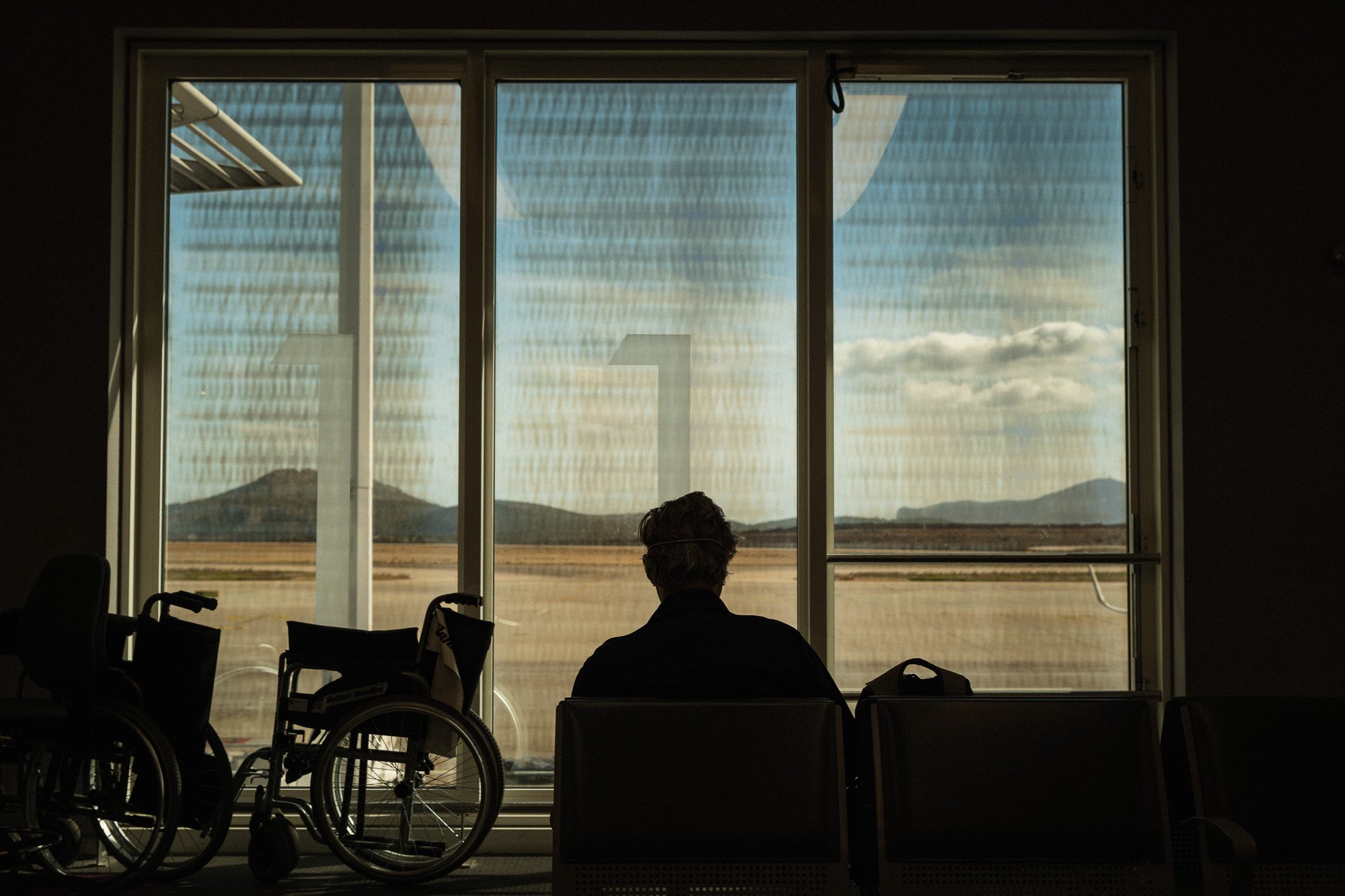 Ein Mensch sitzt in einem Flughafen-Wartesaal neben zwei Rollstühlen vor einem großen Fenster, das eine ländliche Landschaft mit Bergen im Hintergrund zeigt.