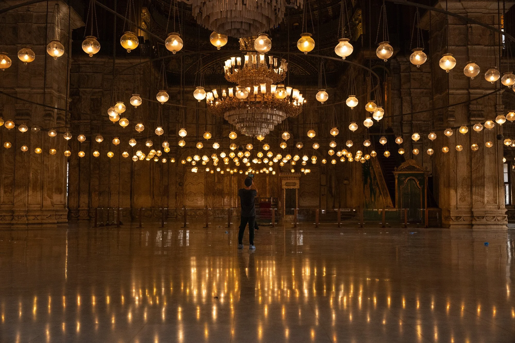 Ein leerer Saal mit zahlreichen hängenden goldfarbenen Lampen und einem großen Kronleuchter in der Mitte. Ein einzelner Mensch steht in der Mitte und fotografiert die Lampen, wölbt sich vor einem Holzwand im Hintergrund.