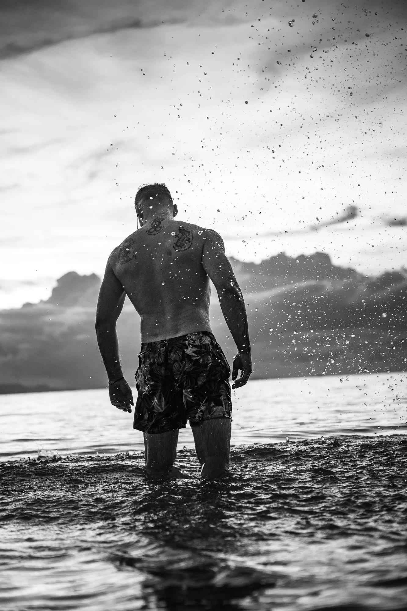 Mann im Wasser, Hinterkörper sichtbar, Sonnenuntergang im Hintergrund, Wasser spritzt, schwarz-weiß Bild.