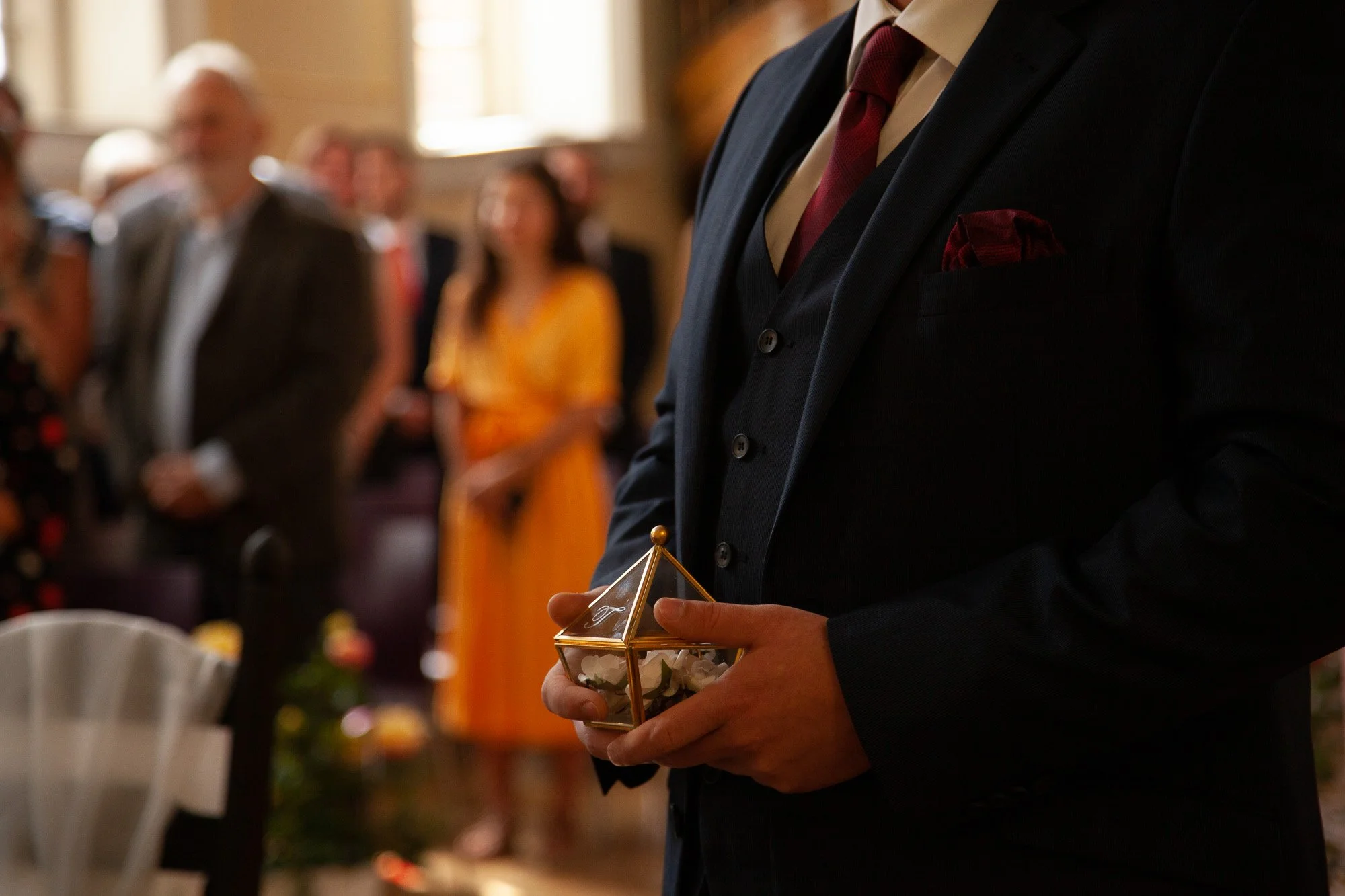 Ein Mann trägt einen dunklen Anzug mit roter Krawatte und hält eine kleine dekorative Glasbox mit Blumen in den Händen bei einer besonderen Veranstaltung, während im Hintergrund Menschen im formellen Kleid in einem Raum mit natürlichem Licht stehen.
