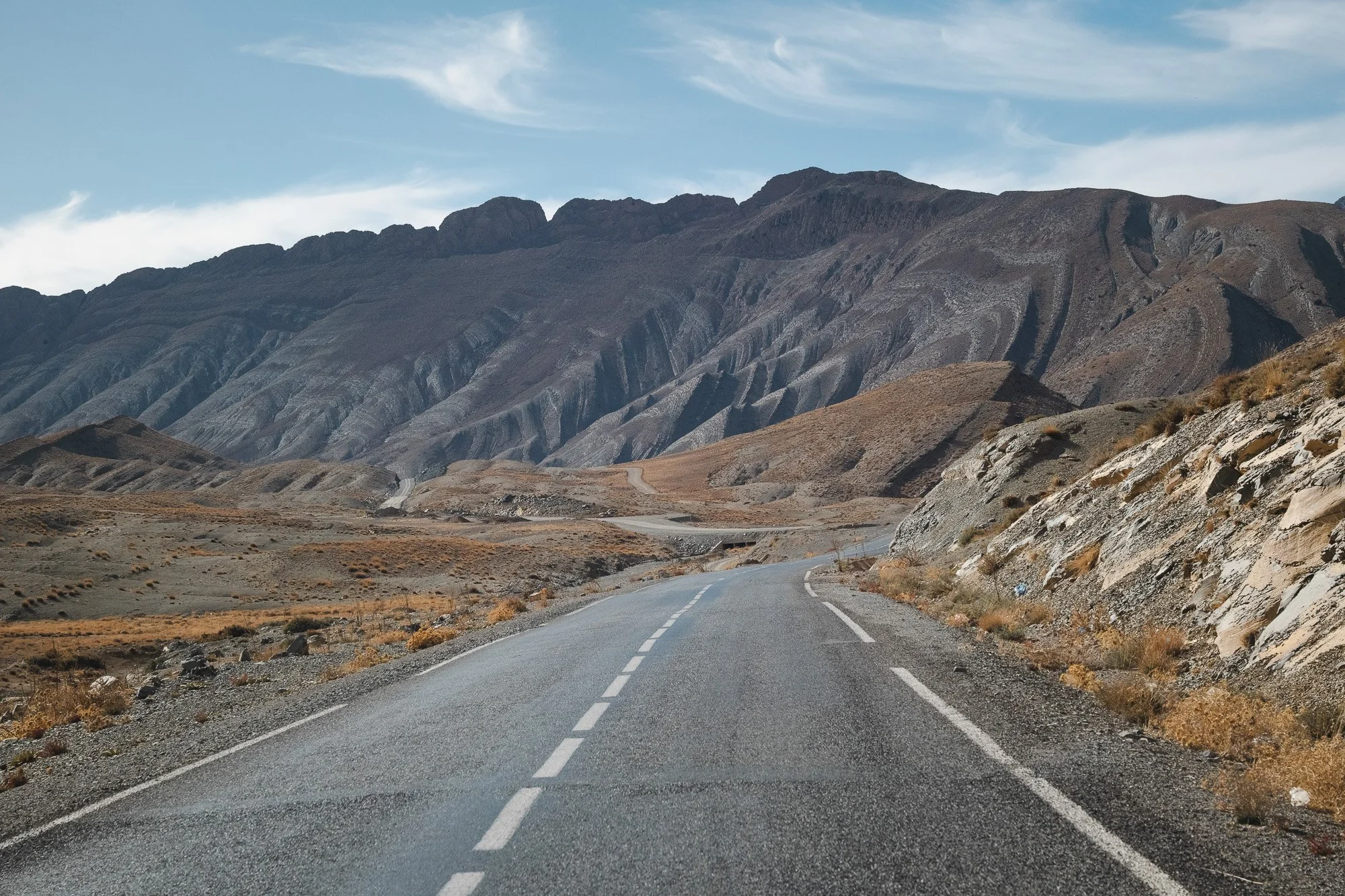Leere Straße in einer bergigen, kargen Landschaft mit schroffen Bergen im Hintergrund und nur wenig Vegetation am Straßenrand.