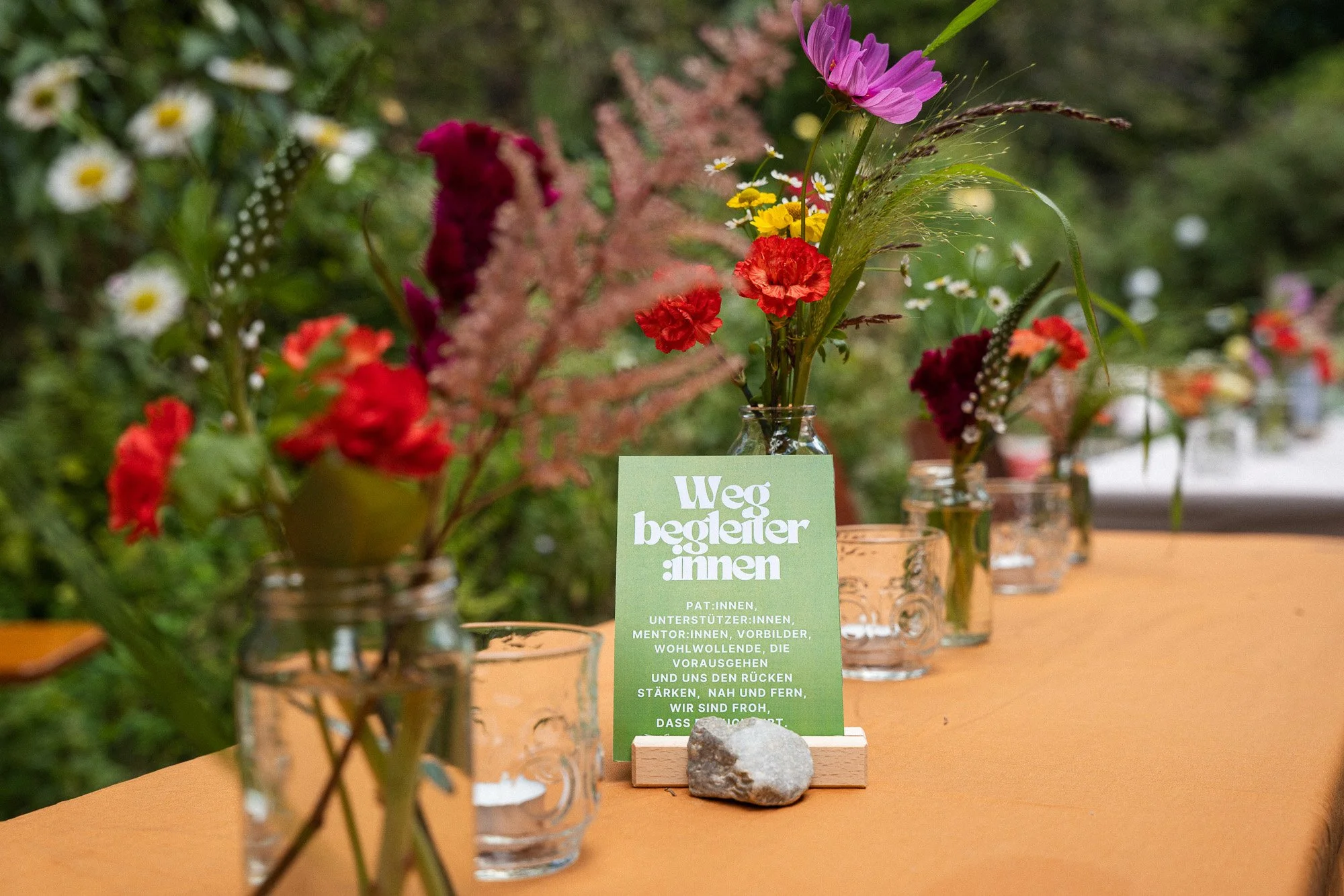 Blumenarrangements in Gläsern auf einem Tisch, mit einer grünen Karte in der Mitte, die einen deutschen Text enthält.