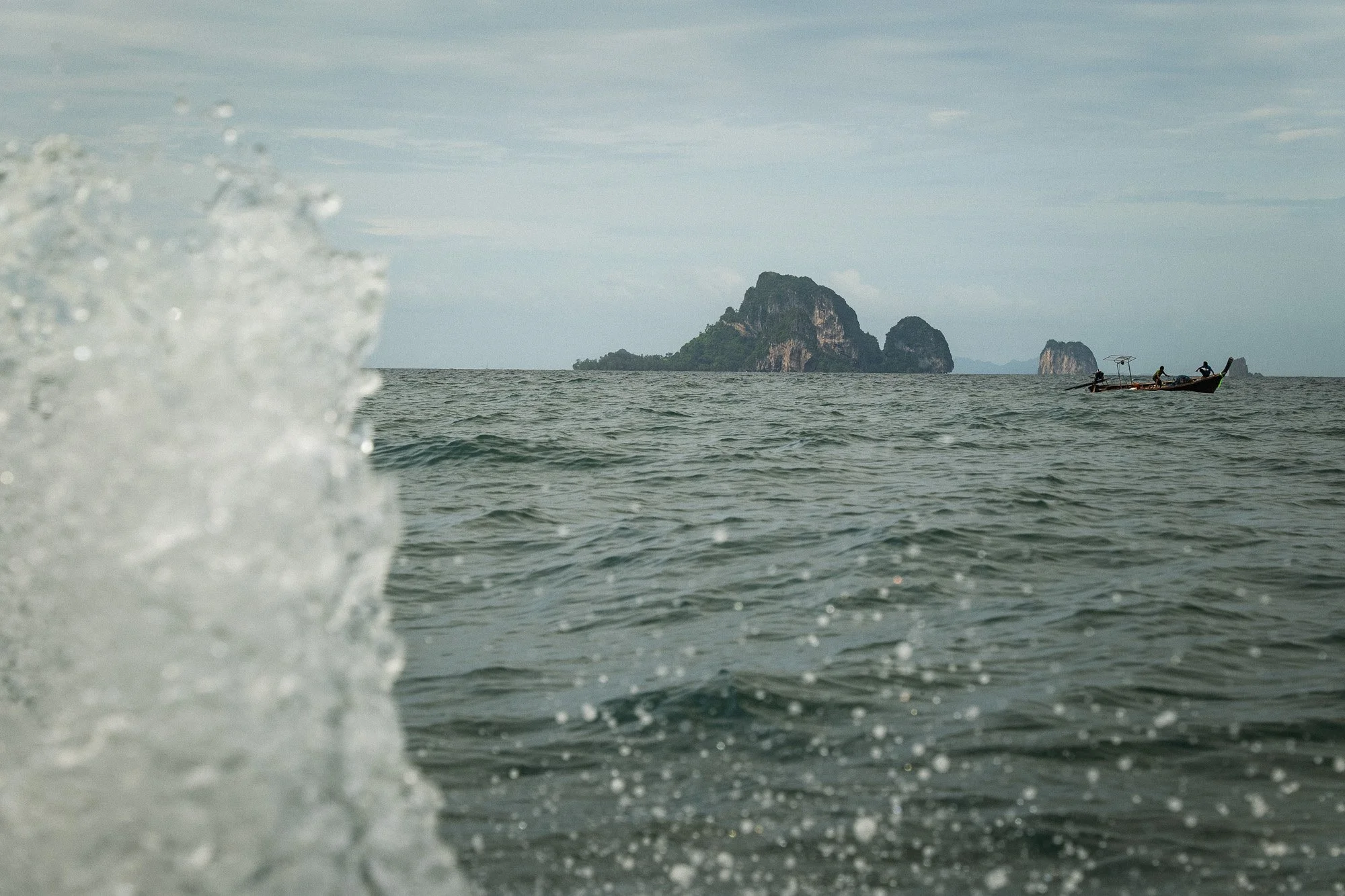 Blick auf das Meer mit einer Welle im Vordergrund, eine kleine Holzkahn mit mehreren Personen auf dem Wasser und eine Insel im Hintergrund.