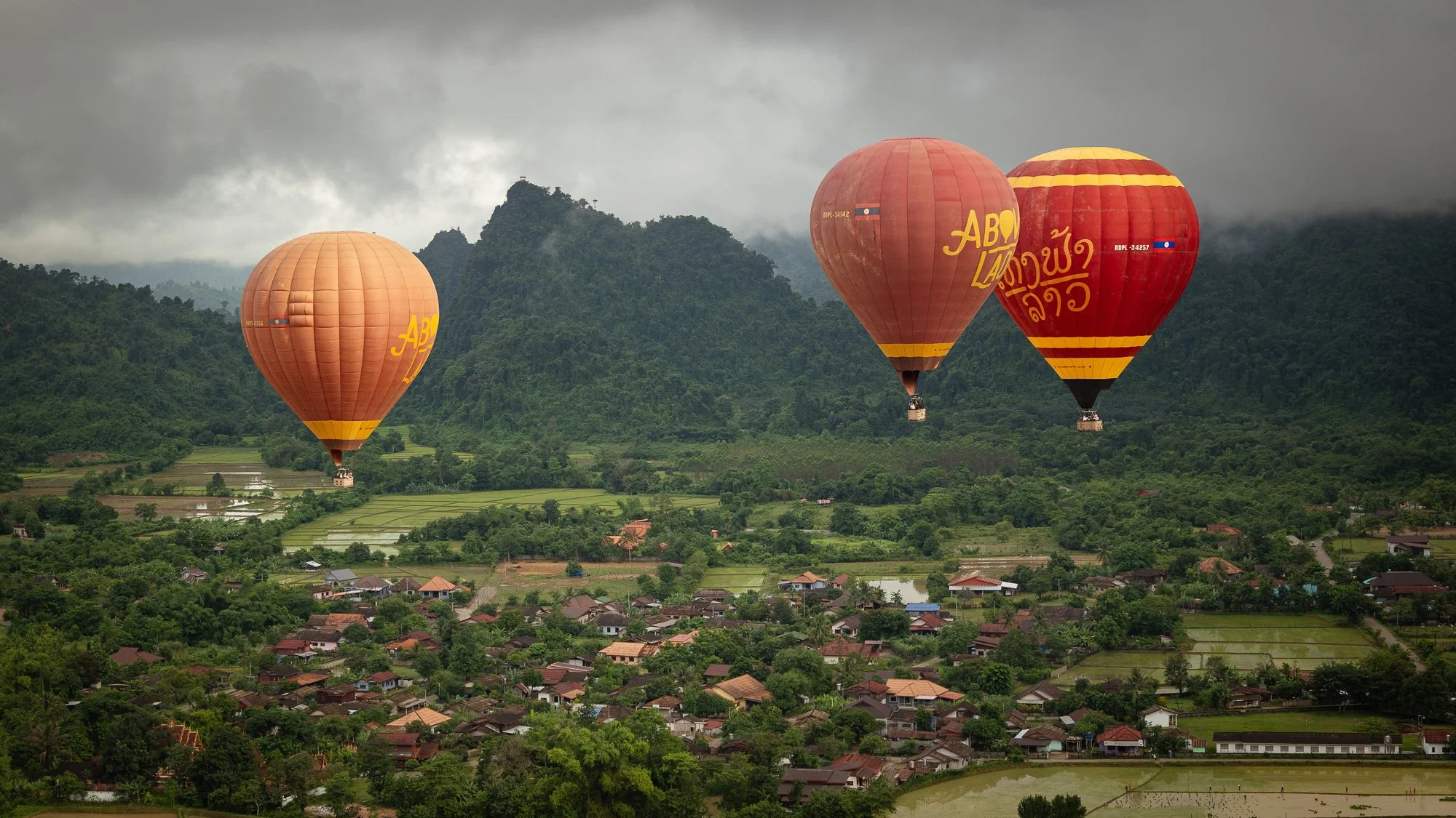 Drei Heißluftballons fliegen über eine grüne Landschaft mit Wohnhäusern, Feldern und Bergen im Hintergrund, bei bewölktem Himmel.