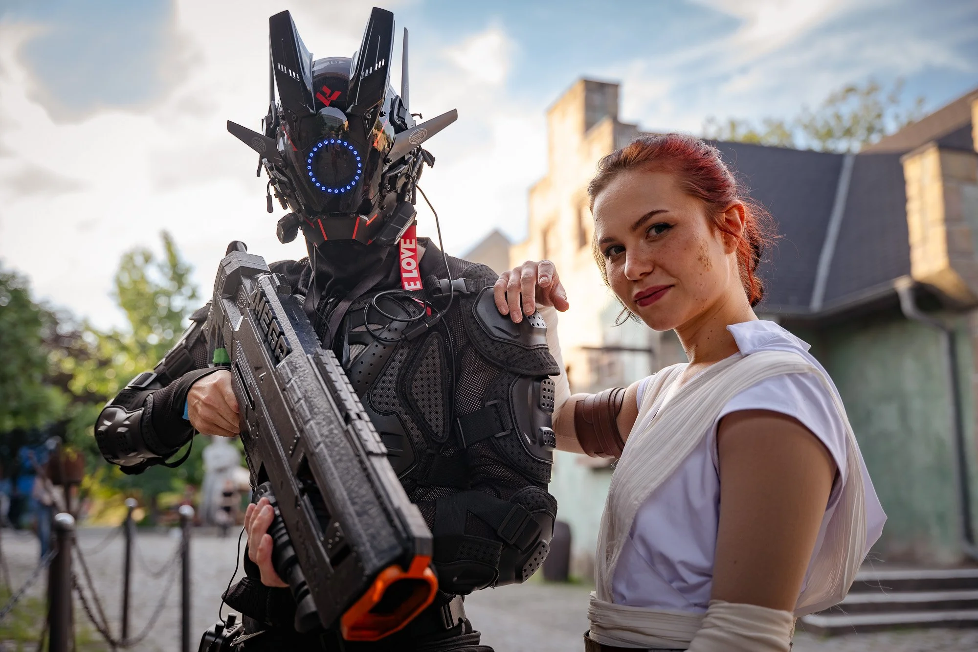 Eine junge Frau mit roten Haaren und einem weißen Kleid, die neben einem humanoiden Roboter steht, der einen futuristischen Helm trägt und eine große Waffe hält, vor einer alten Gebäude- und Baumkulisse bei Tageslicht.