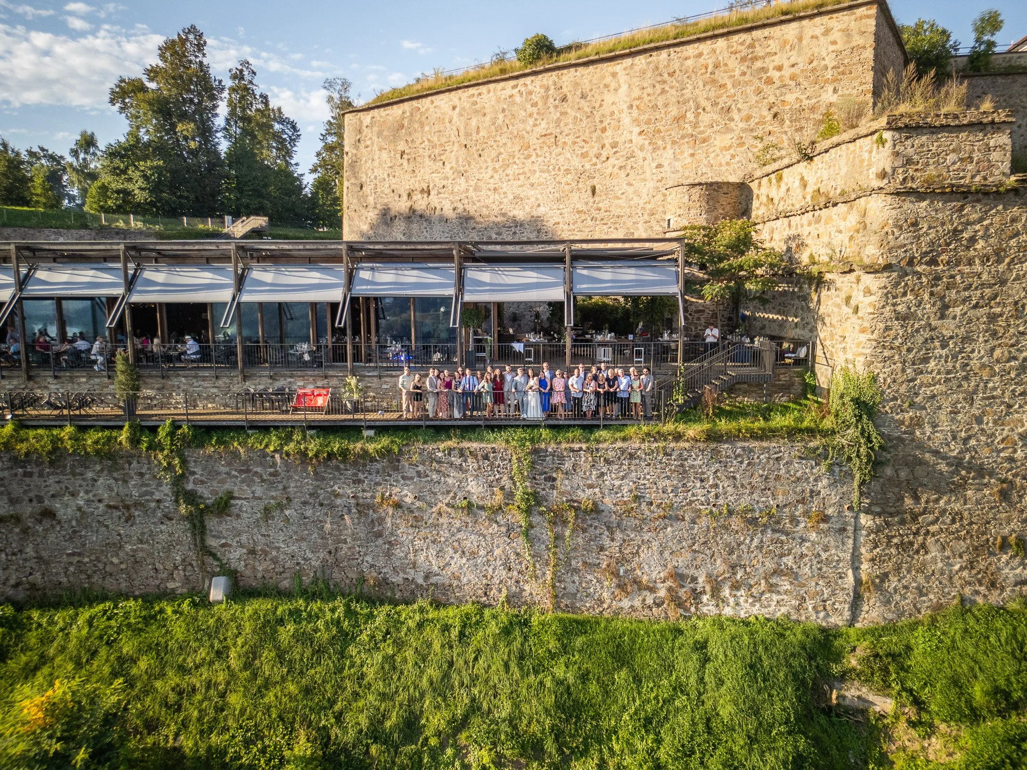Eine Gruppe von Menschen steht auf einer Terrasse vor einer alten Steinmauer mit Blick auf einen Garten. Es ist Tag mit bewölktem Himmel.