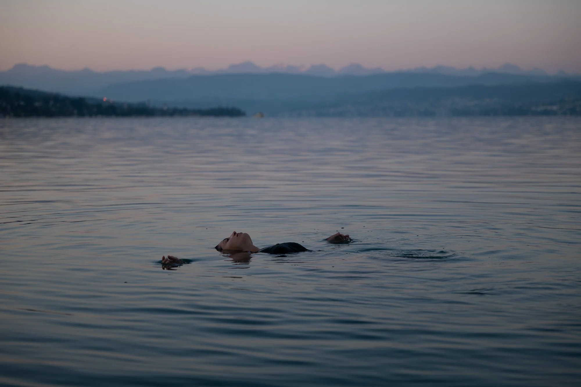 Eine Person liegt auf dem Rücken im Wasser eines Sees, mit geschlossenen Augen, bei Sonnenuntergang, mit Bergen im Hintergrund.