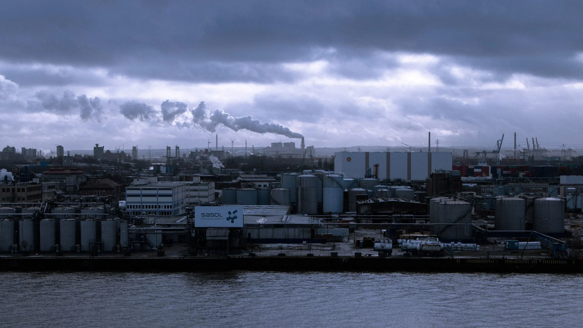 Industriehafen mit Fabriken, Rohre und Frachtcontainer, dunkler Himmel, Rauch und Dampf steigen aus den Schornsteinen, Wasser im Vordergrund, dunkle Wolken am Himmel.