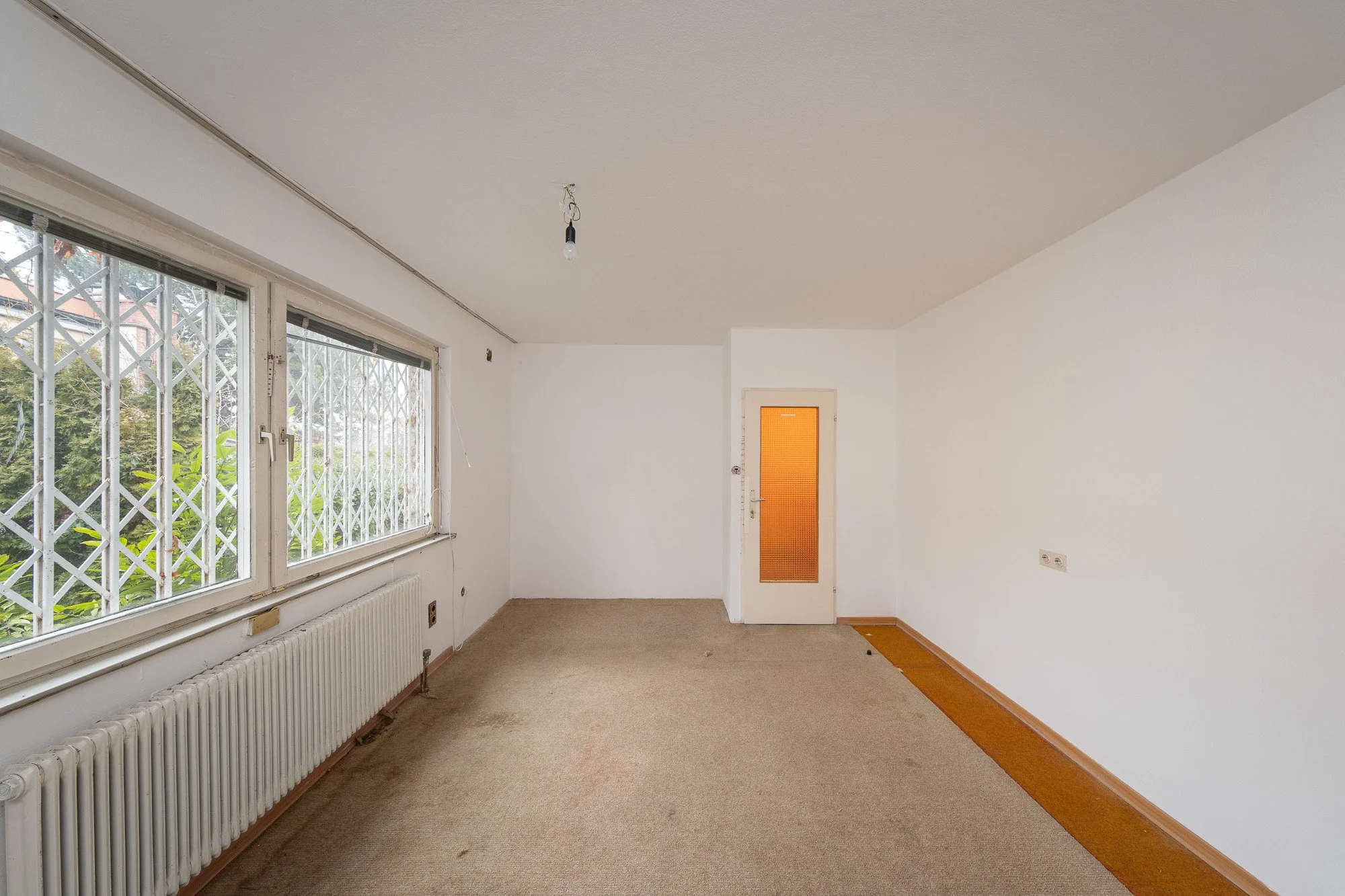 leerer Raum mit weißen Wänden, einem braunen Teppichboden und Fenster mit Sicherheitsgittern, die auf die Außenseite blicken, eine Tür mit orangener Glasfüllung, keine Möbel, Kabel an der Decke, Heizkörper entlang der Wand unter den Fenstern