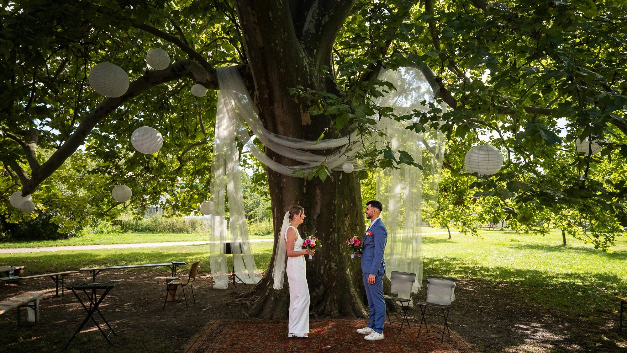 Ein Brautpaar steht unter einem Baum bei ihrer standesamtlichen Trauung im Freien, mit weißen Papierlaternen und Dekorationen wie Tüll unter den Ästen.