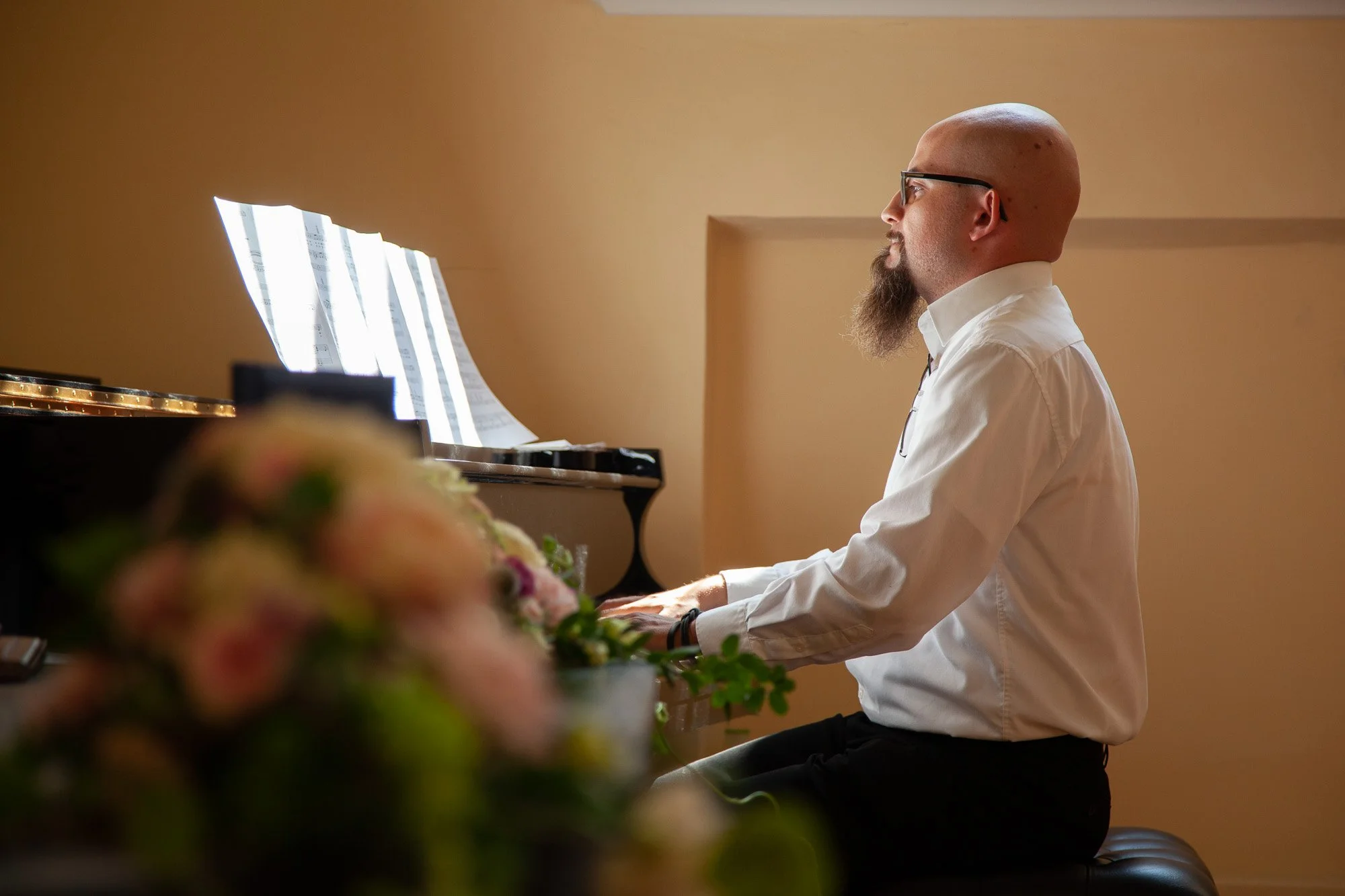 Mann spielt Klavier im Raum, vor ihm Blumen, während er sich auf das Spielen konzentriert.