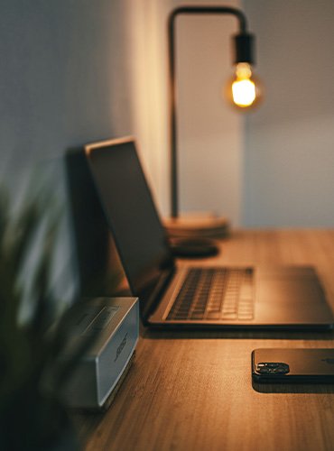 Ein Arbeitszimmer mit einem offenen Laptop, einem Smartphone und einem Tischlampen in warmem Licht.