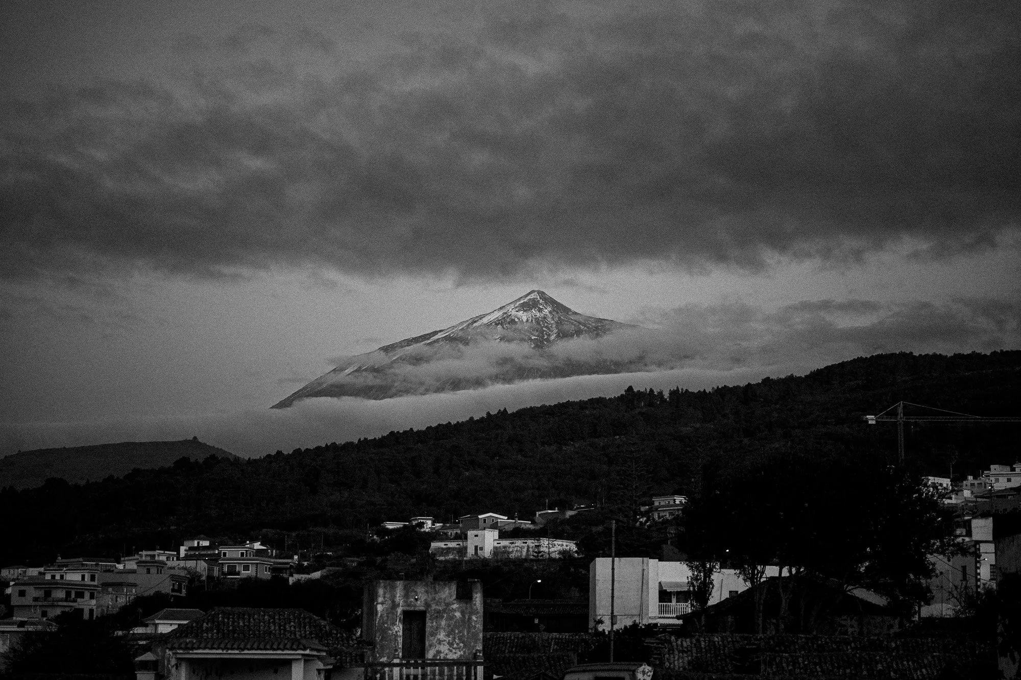 Schwarz-weiß-Foto eines Vulkans mit Schnee auf dem Gipfel, über einer Stadt mit Häusern und Bäumen, Wolken hängen tief über dem Vulkan.