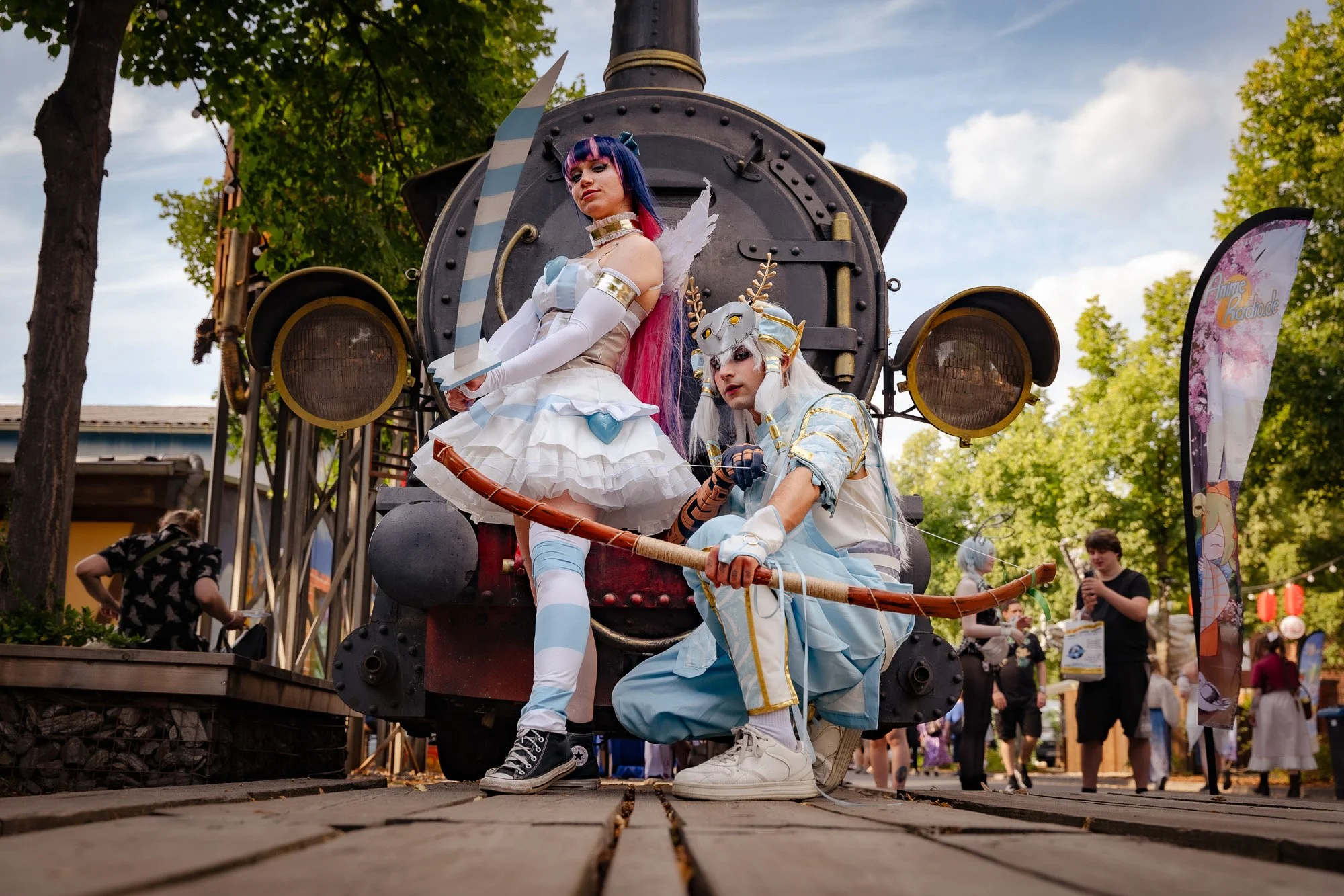 Zwei Cosplayer in Fantasy-Kostümen posieren vor einem alten Dampfzug bei einem Outdoor-Event. Der Hintergrund zeigt Bäume, einen blauen Himmel und weitere Besucher.