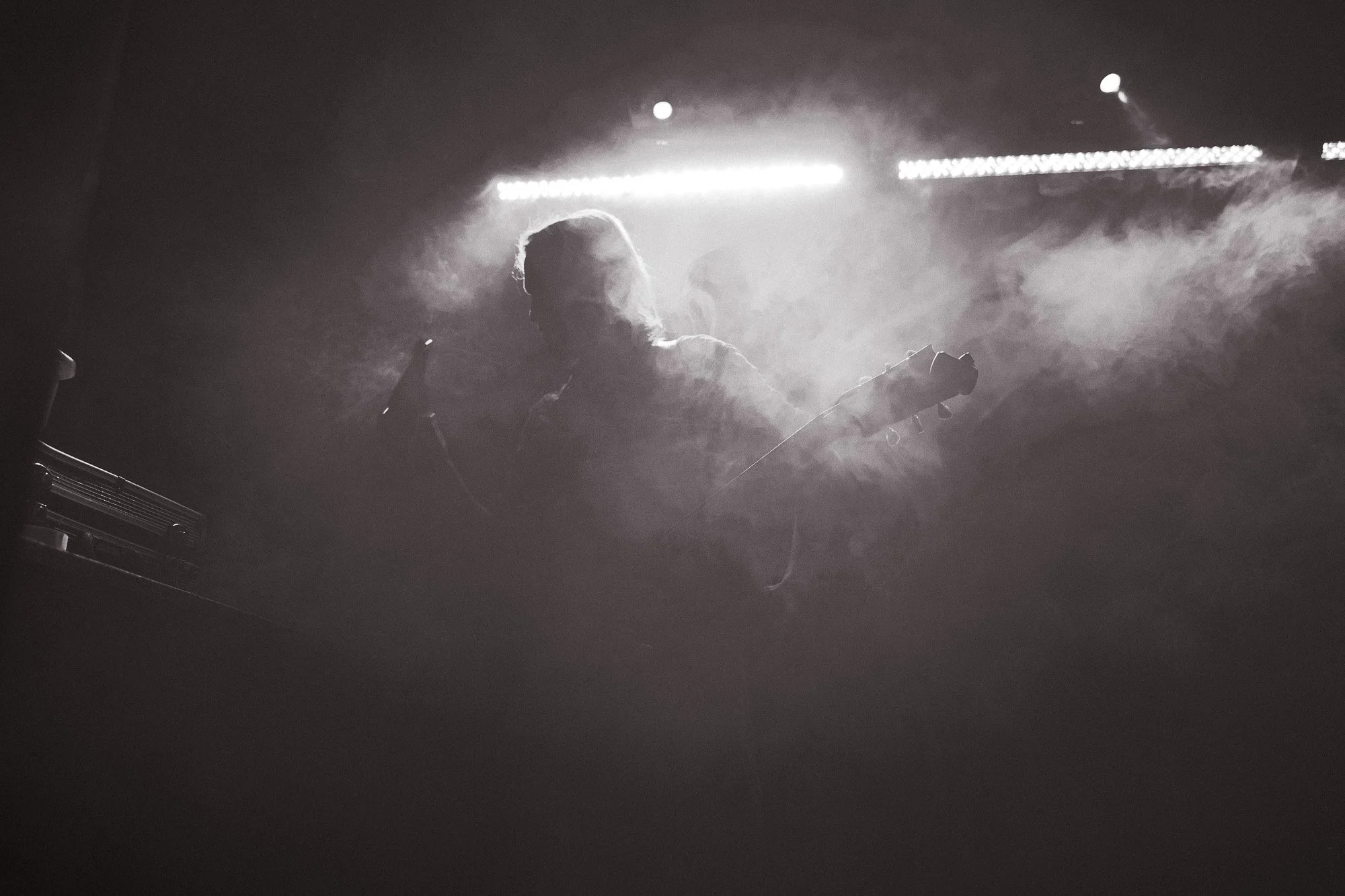 Eine dunkle Szene mit einem Musiker, der eine Gitarre hält, im Nebel und bei schwachem Licht auf einer Bühne.