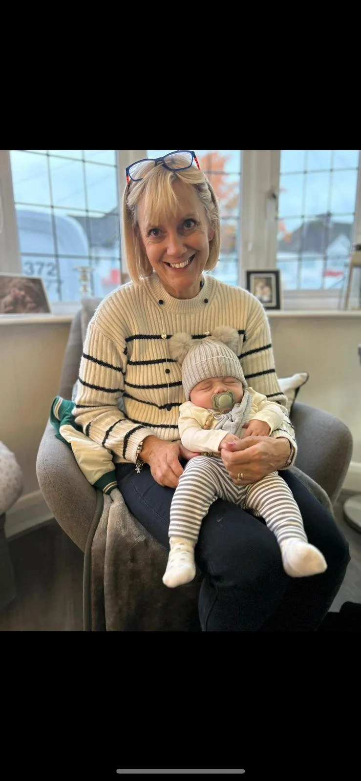 A woman smiling and holding a disposable coffee cup and a brown folder, standing indoors with plants and large windows in the background.