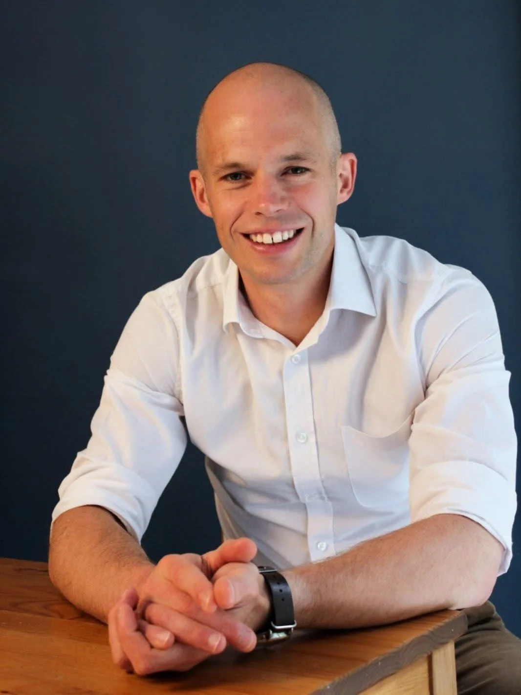A smiling man with a shaved head wearing a white button-up shirt with rolled-up sleeves, sitting at a wooden table against a dark blue background.