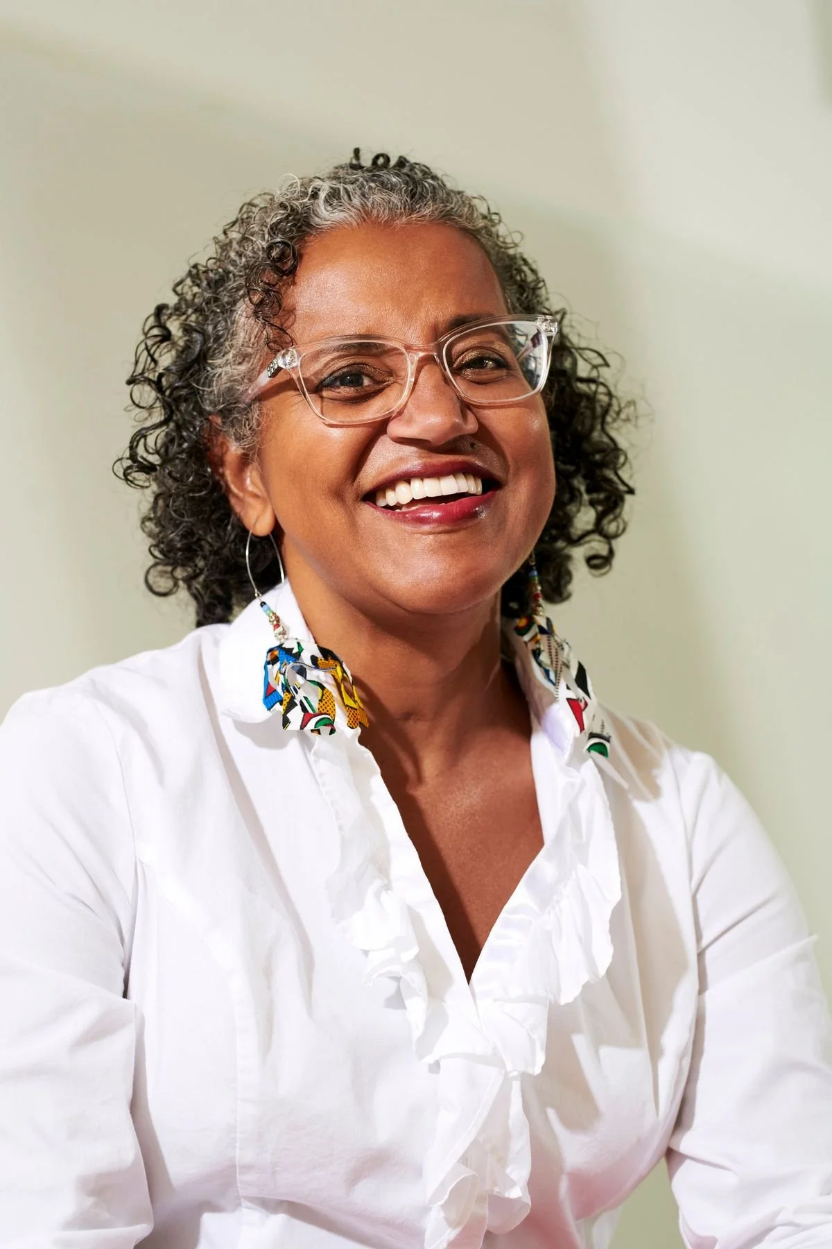 A smiling woman with curly gray hair wearing glasses, colorful earrings, and a white ruffled blouse.